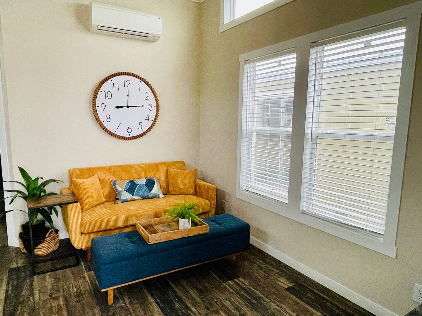 Cozy living room with mustard sofa, blue ottoman, and a round wall clock. Large windows with blinds let in natural light. Warm, inviting decor.