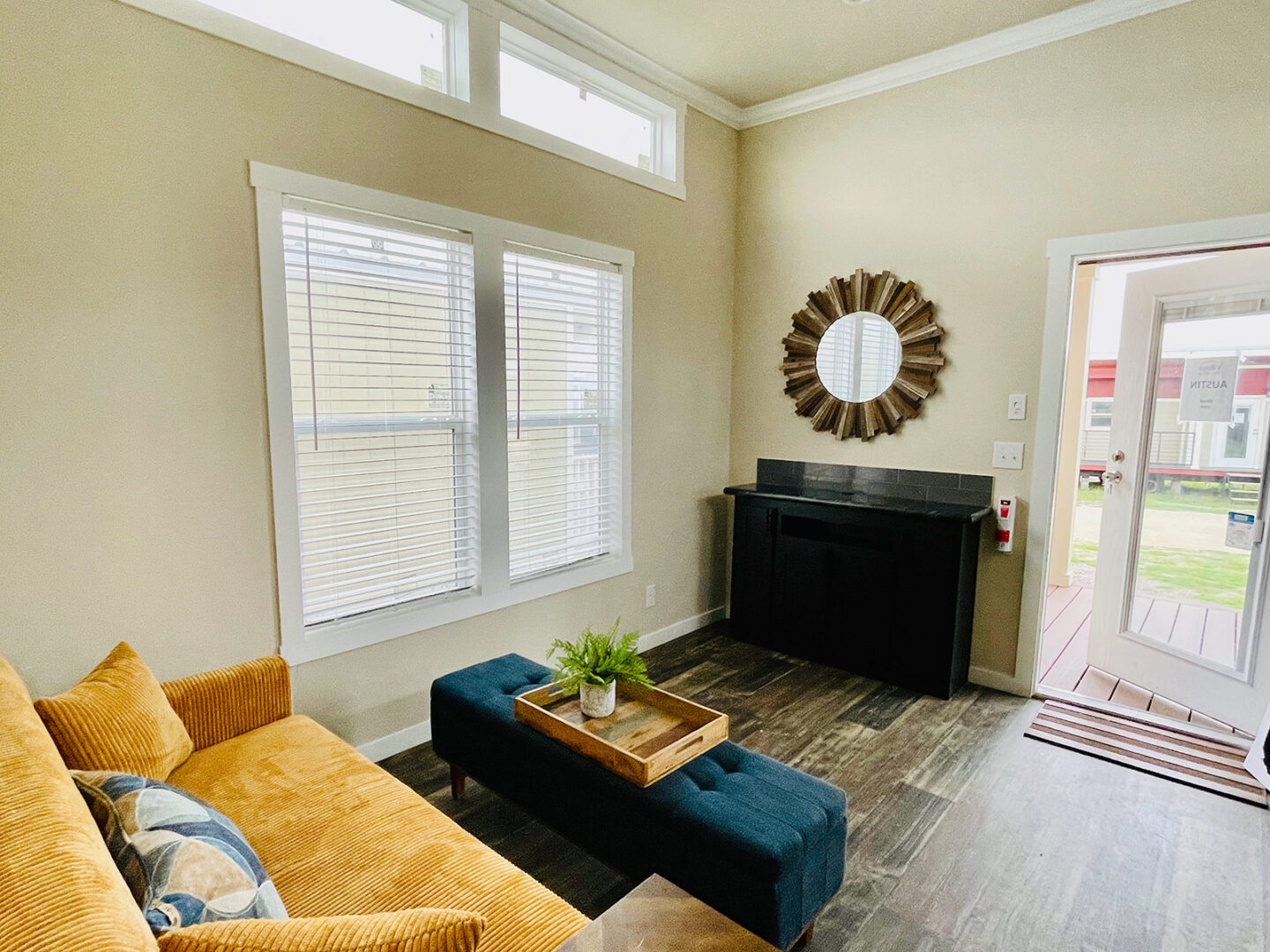 Cozy living room with a golden couch and patterned pillows, a dark ottoman with a wooden tray and plant. Sunburst mirror and large windows brighten space.