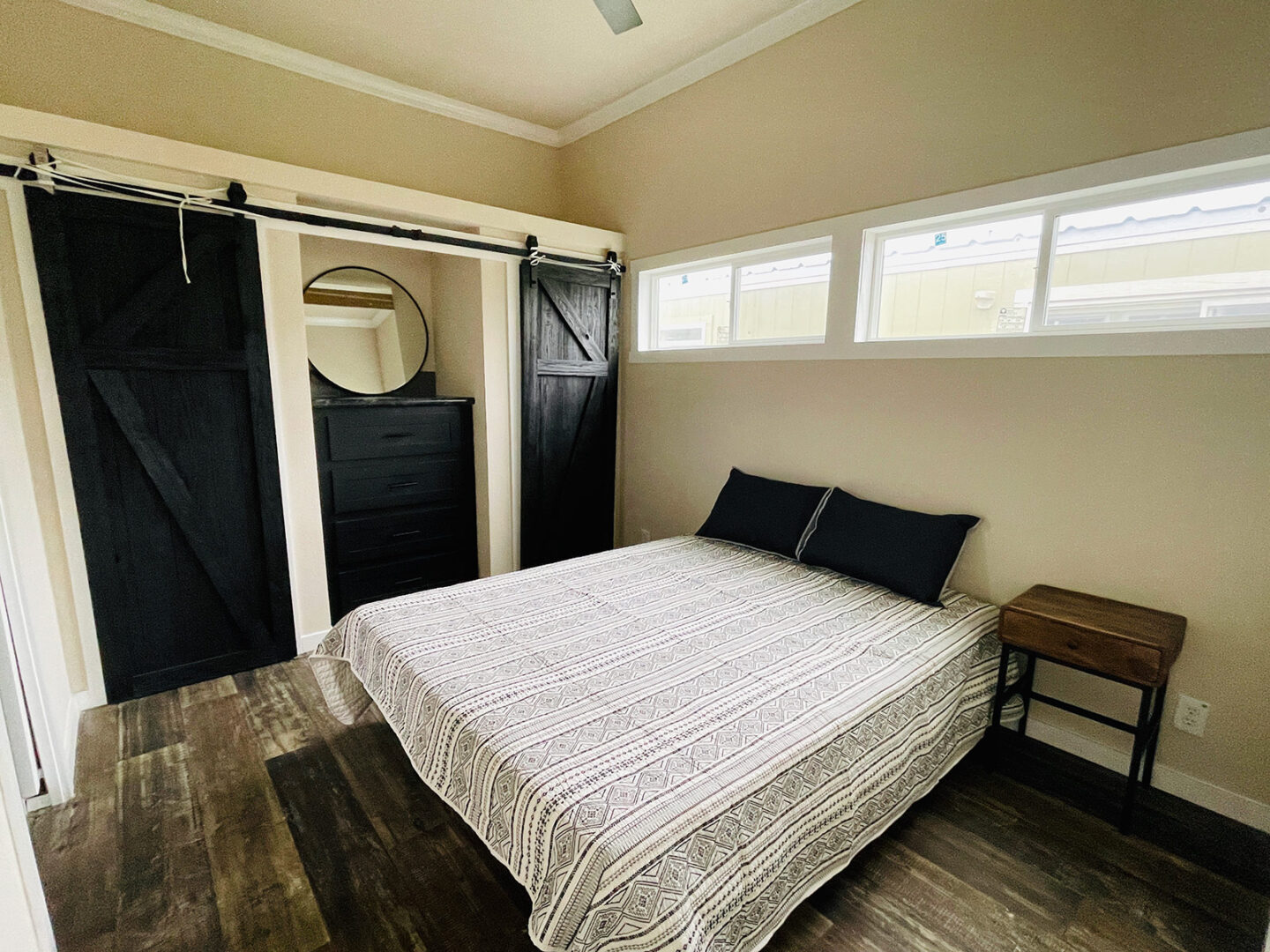 Cozy bedroom with a rustic vibe. Features dark wood sliding barn doors, a bed with patterned bedding, a round mirror, and a wooden nightstand.