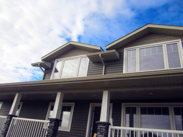 Two-story house with gray siding, white trim, and large windows. A covered porch with white railings and stone columns. Partly cloudy sky above.