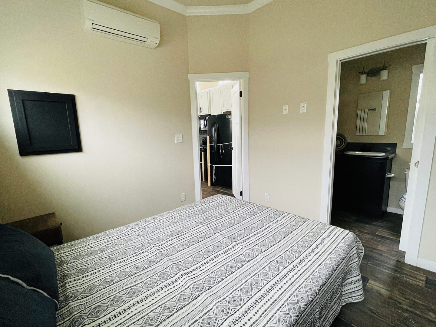 A cozy bedroom with a patterned bedspread. Doors lead to a kitchen with a black fridge and a bathroom. Warm tones evoke a calm, inviting atmosphere.