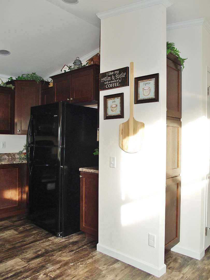 A cozy kitchen with warm wooden cabinets, a black fridge, and wood-look flooring. Wall decor includes coffee-themed art and a wooden pizza peel.
