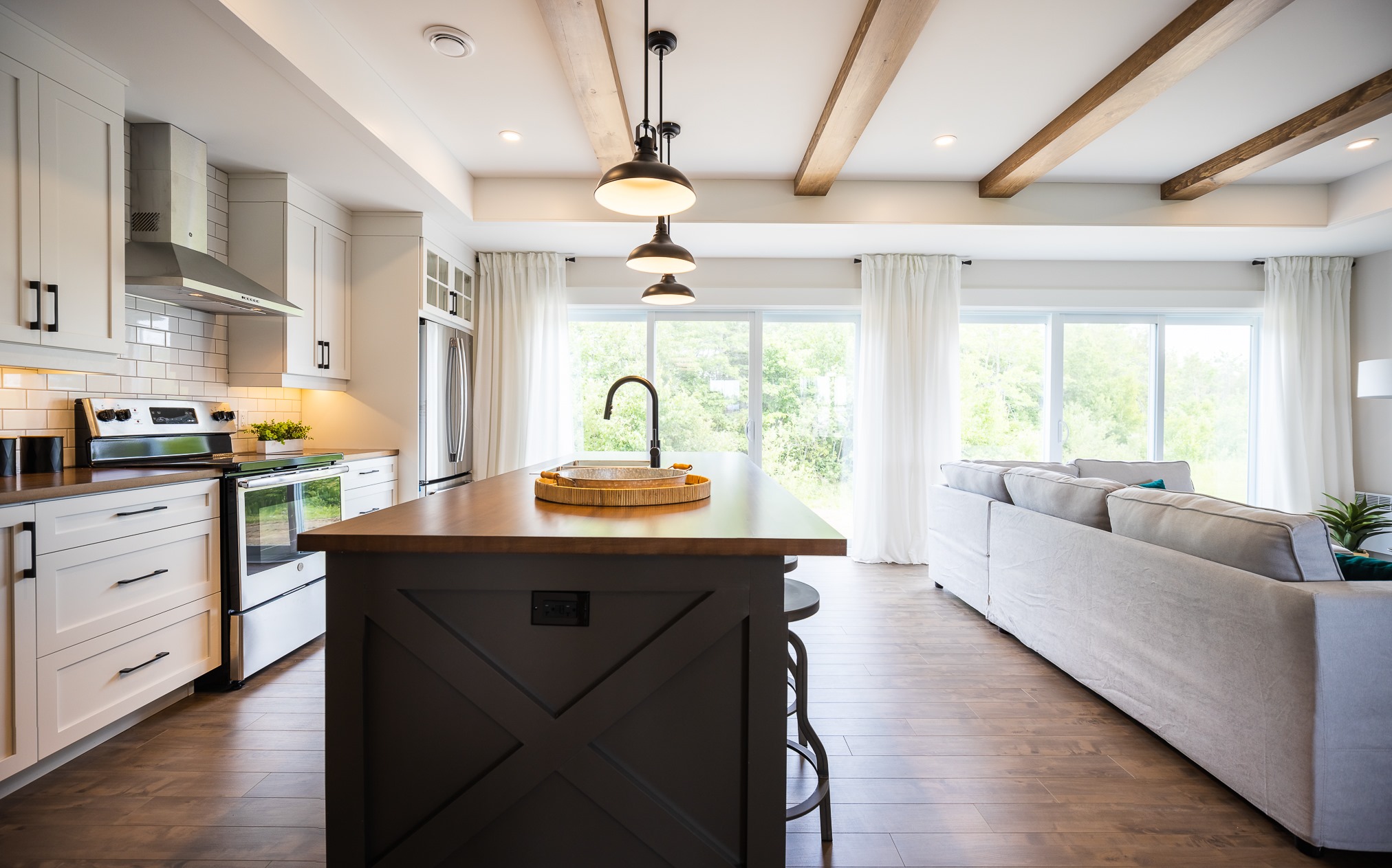 Spacious, bright kitchen and living area with a central island, wood beams, and large windows. Creamy tones and cozy atmosphere with modern decor.