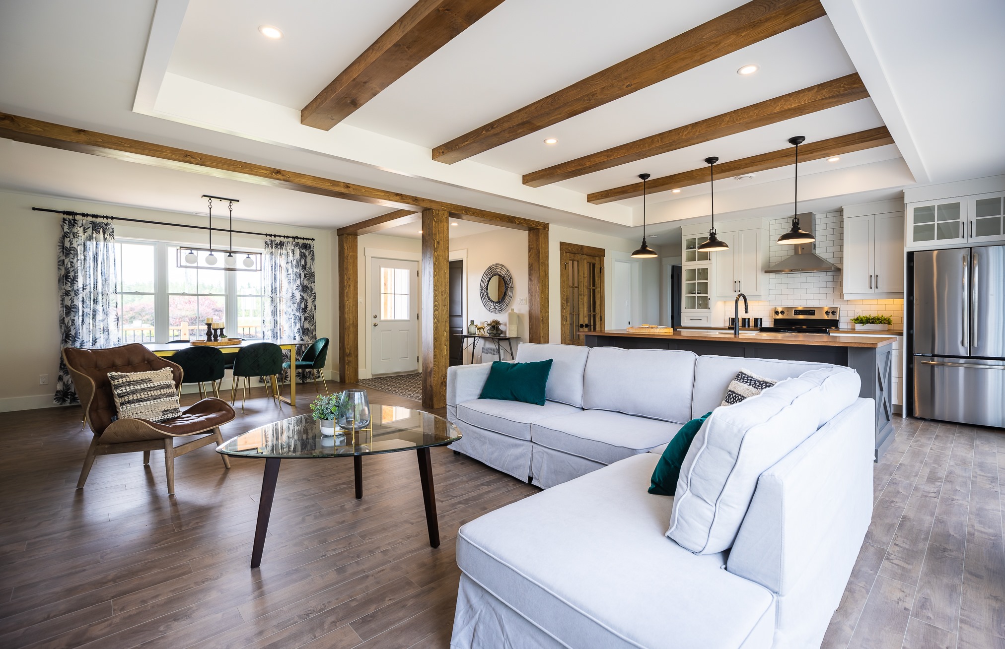 Spacious modern open-concept living room and kitchen with wooden beams, white L-shaped sofa, glass coffee table, and elegant dining area. Relaxing ambiance.