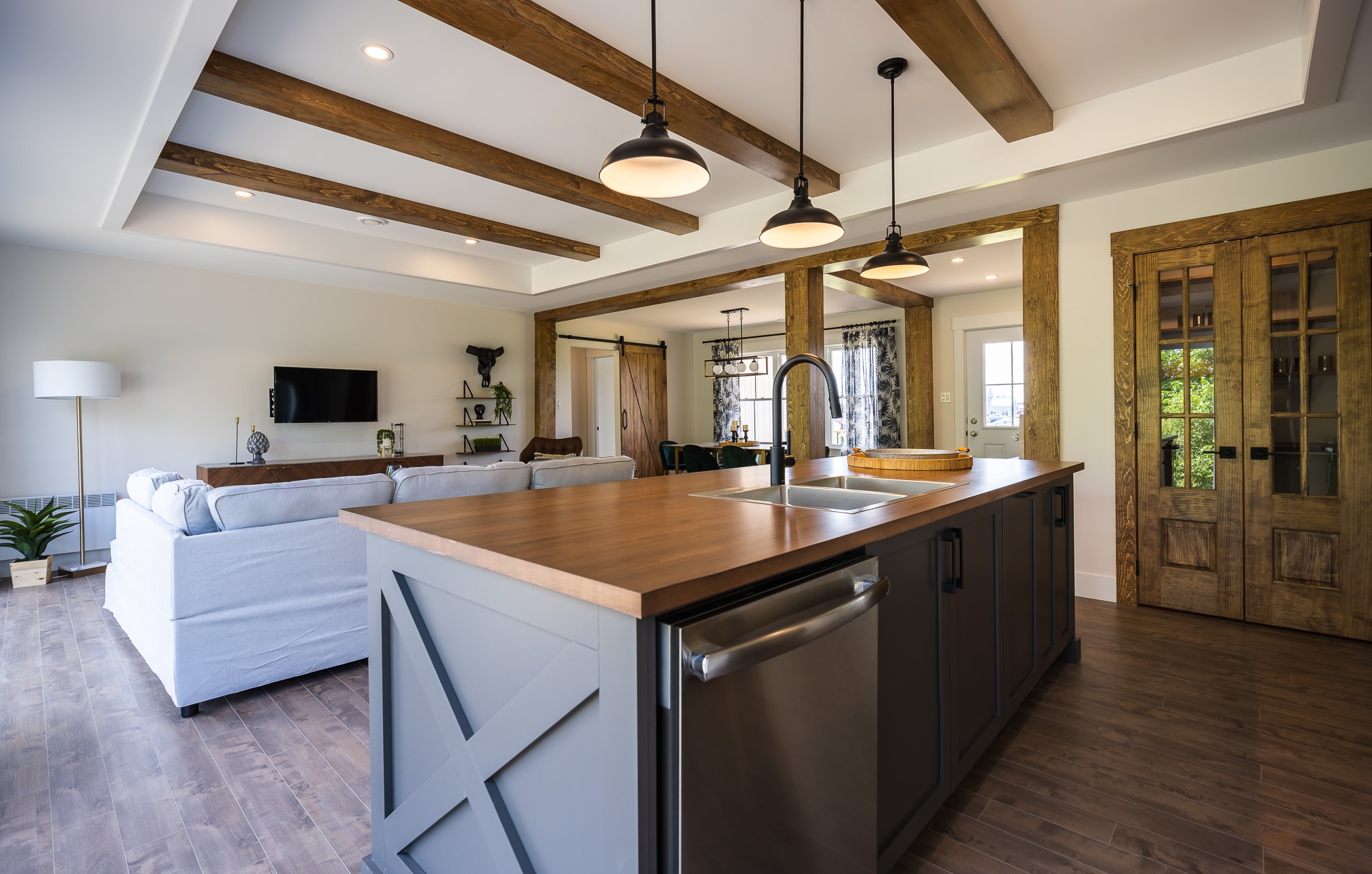 A modern farmhouse kitchen features wooden beams, a large island with a sink, and pendant lights. The open layout extends to a cozy living area.