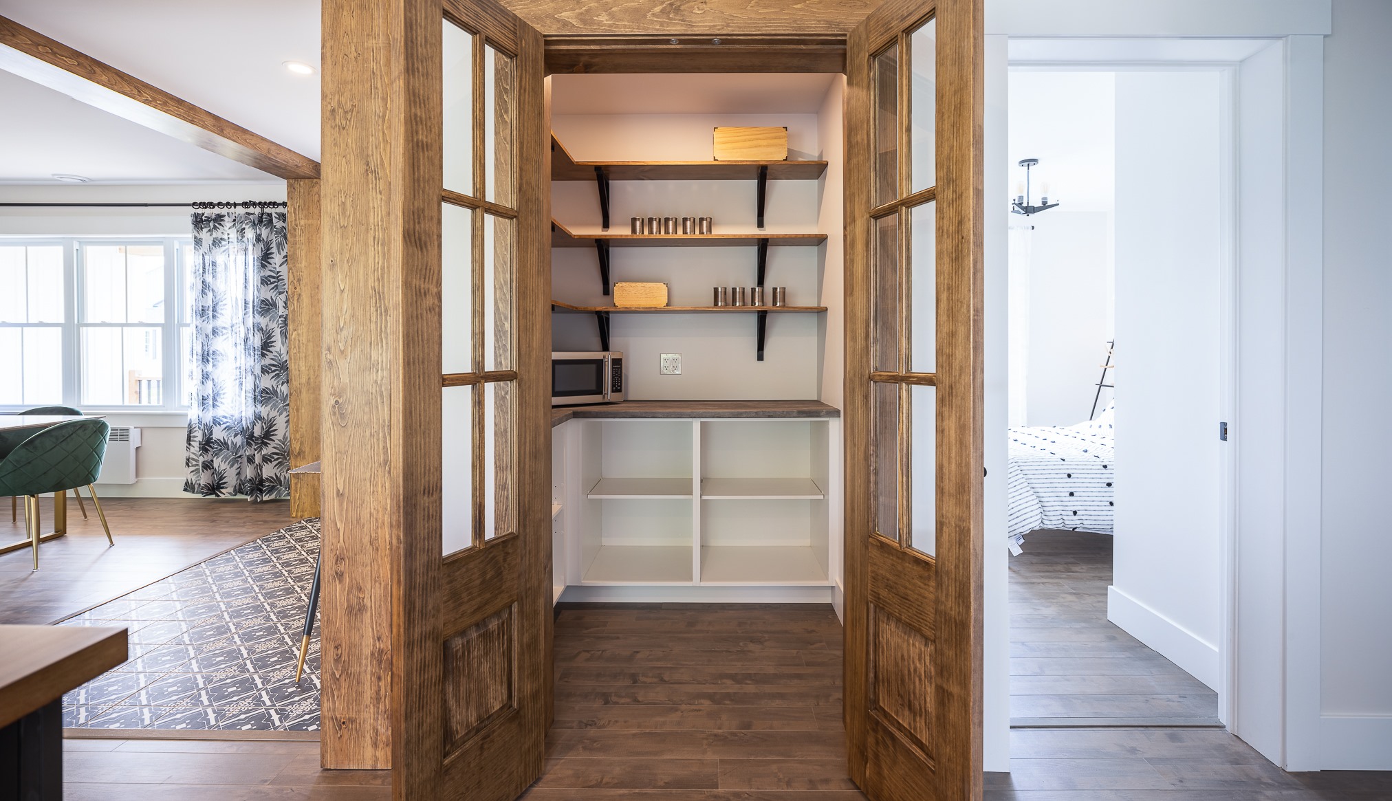 A cozy room features rustic wooden double doors opening to a small pantry with shelves holding a microwave, jars, and linens. Natural light pours in from a large window in the adjacent space, creating a warm, welcoming ambiance.