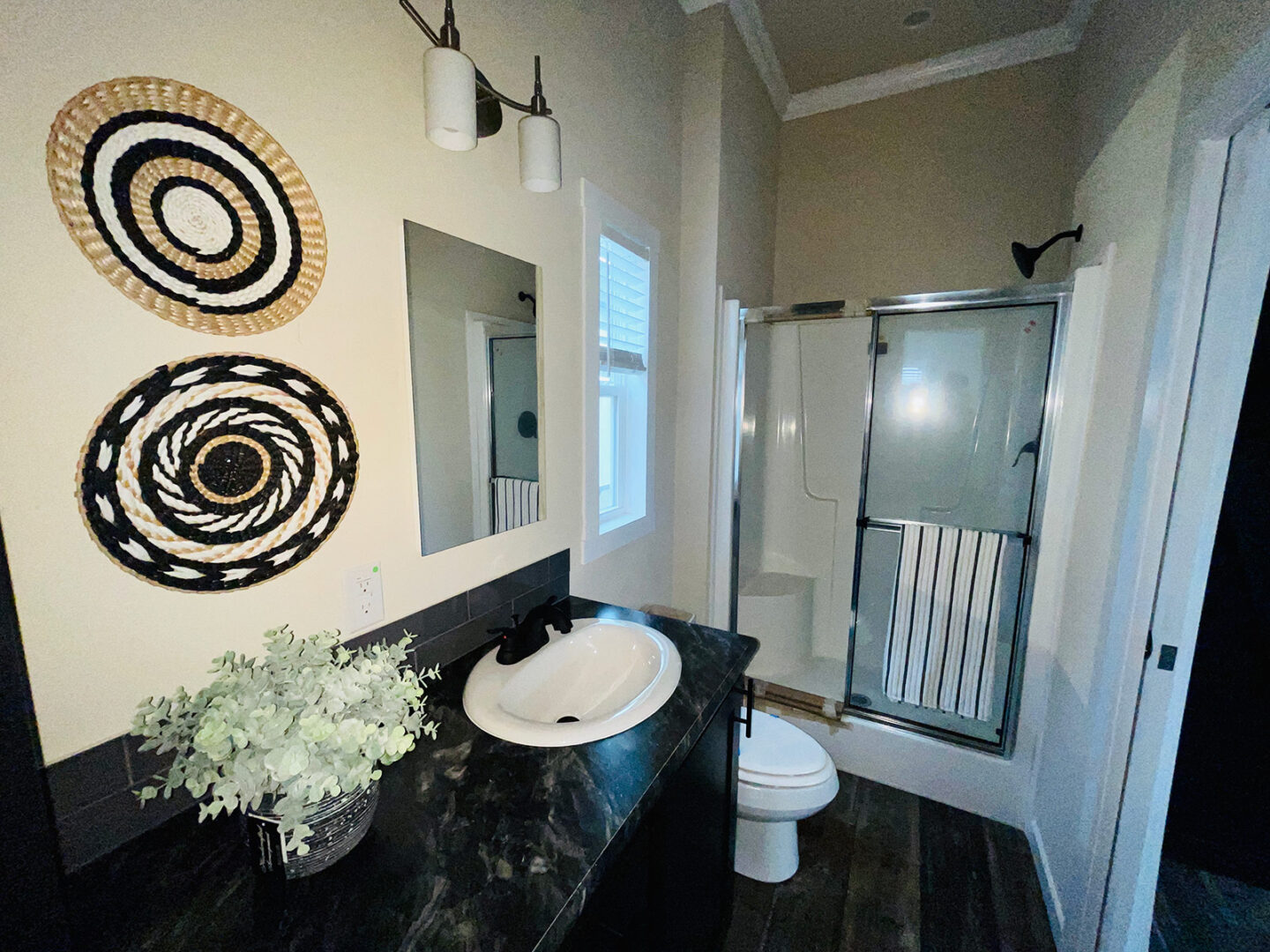 Modern bathroom with dark marble countertop, white sink, and leafy plant. Two woven baskets hang on the wall; a glass shower is in the background.