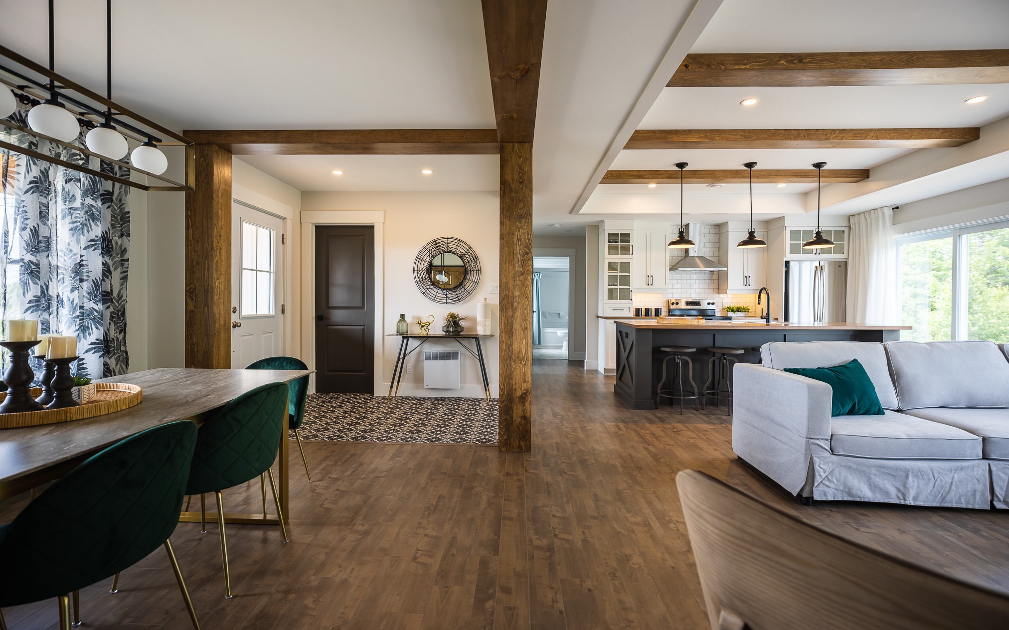 Open-concept living space with wooden floors and beams, featuring a dining area with green chairs, a cozy seating area, and a modern kitchen island.