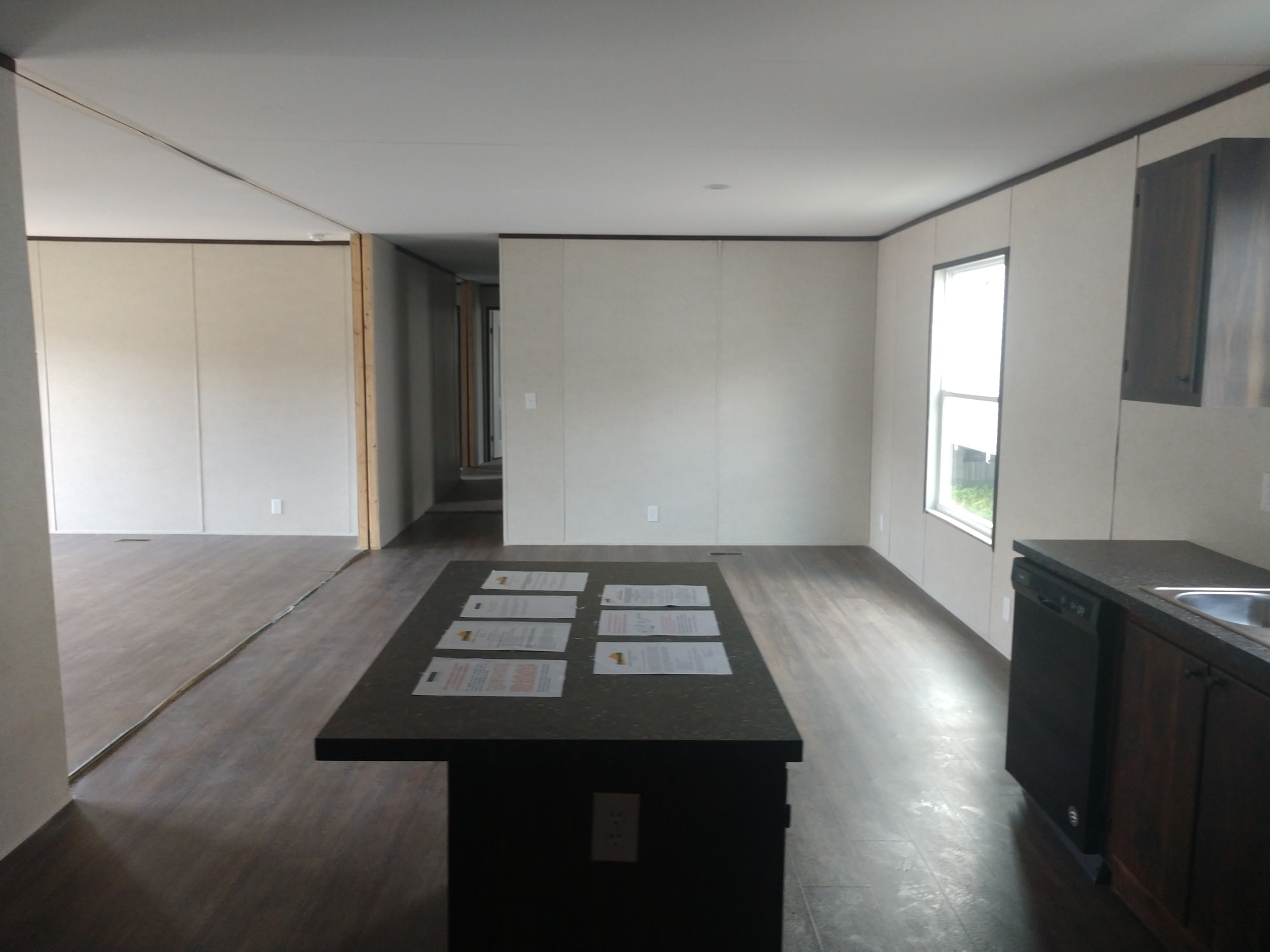 Spacious, unfurnished room with light walls and wood flooring. A kitchen island with papers on it is centered. Natural light fills the room from a window.
