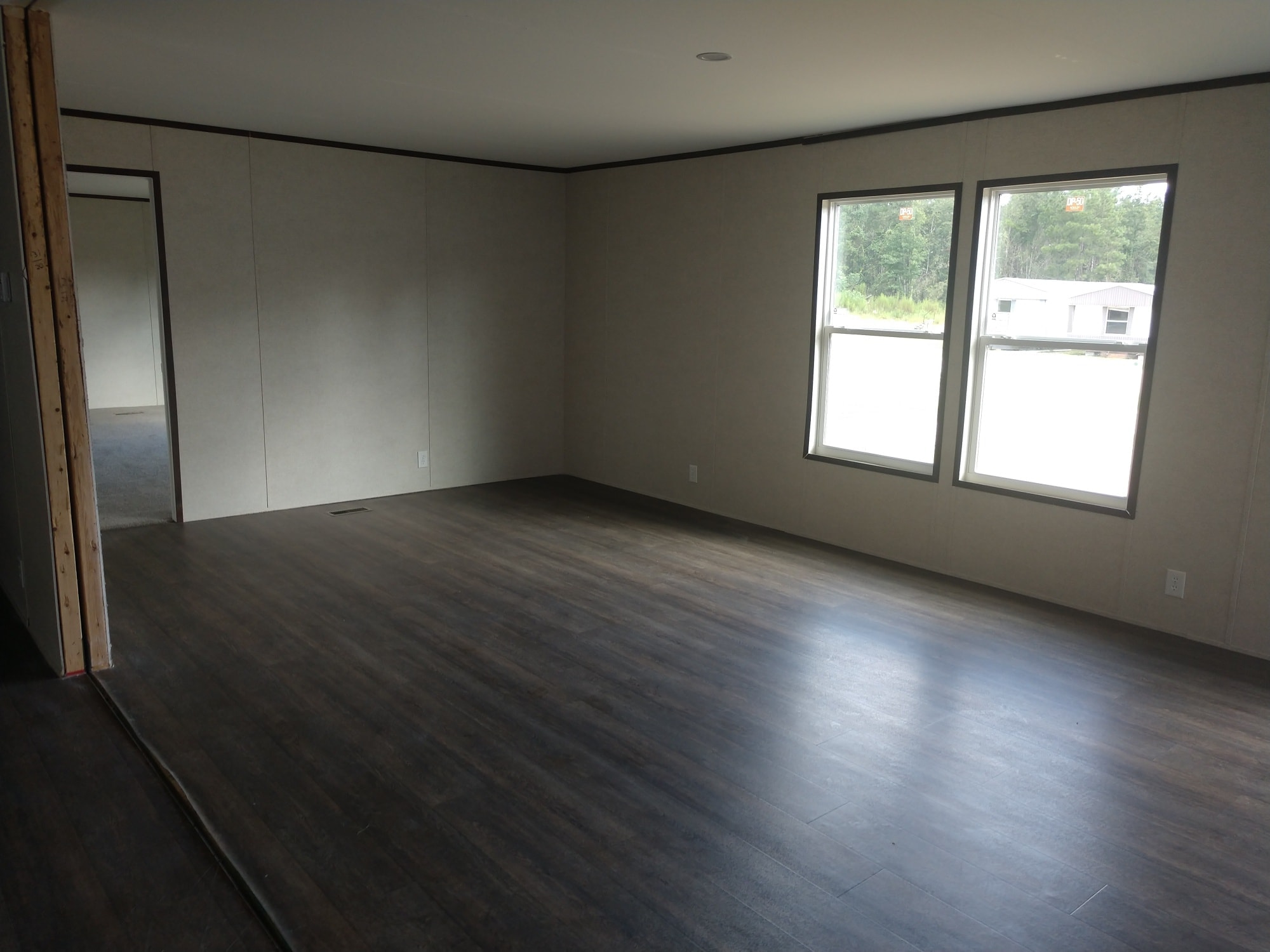 Empty room with dark wood flooring and beige walls. Two windows allow natural light, revealing a doorway on the left. Calm and minimalistic atmosphere.