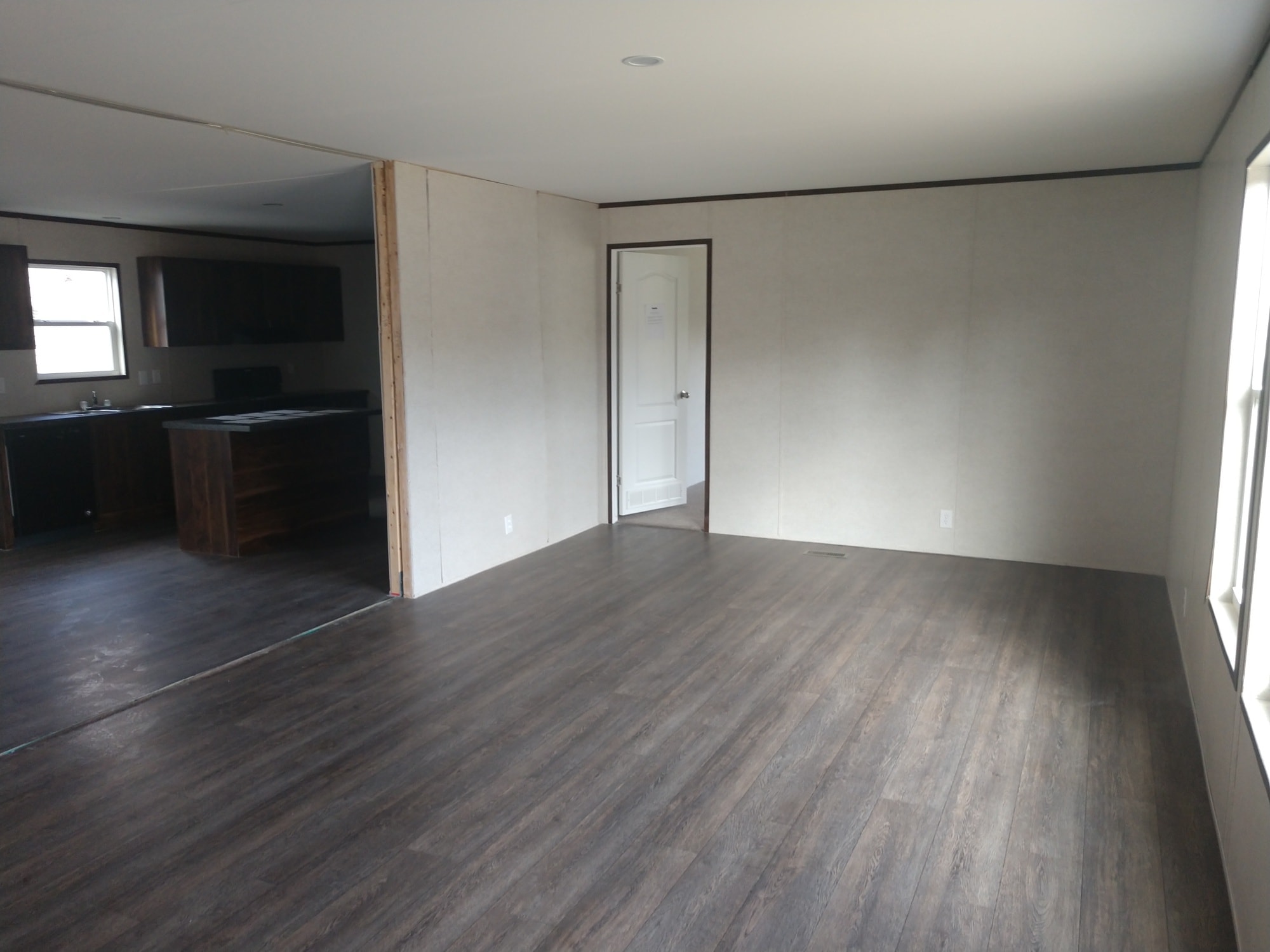 Empty room with dark wooden floors and light gray walls, featuring a white door. Sunlight streams in from a window, creating a calm atmosphere. Kitchen visible to the left.
