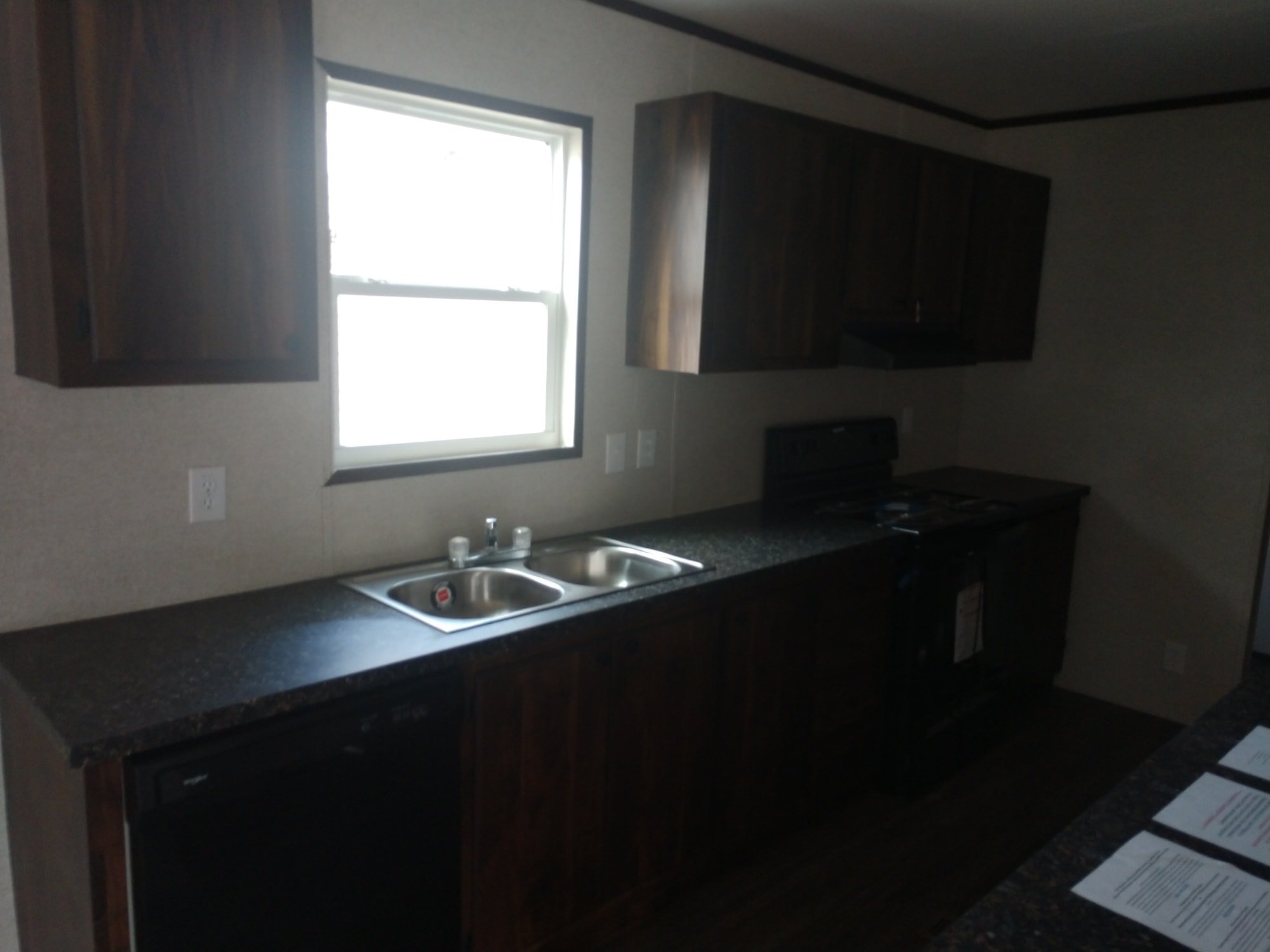 Dark wood kitchen with brown cabinets, a stainless steel sink under a sunny window, and black appliances. A dimly lit, minimalist design.