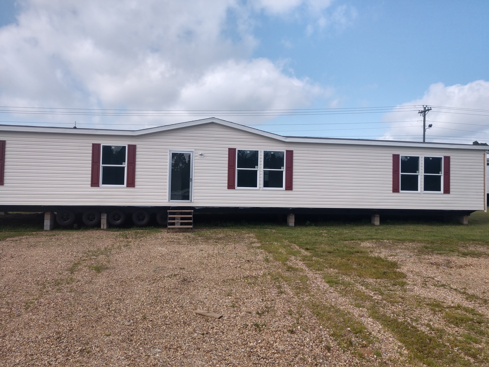 A single-story mobile home with a white exterior and red shutters sits on a gravel lot under a cloudy blue sky, conveying a calm rural setting.