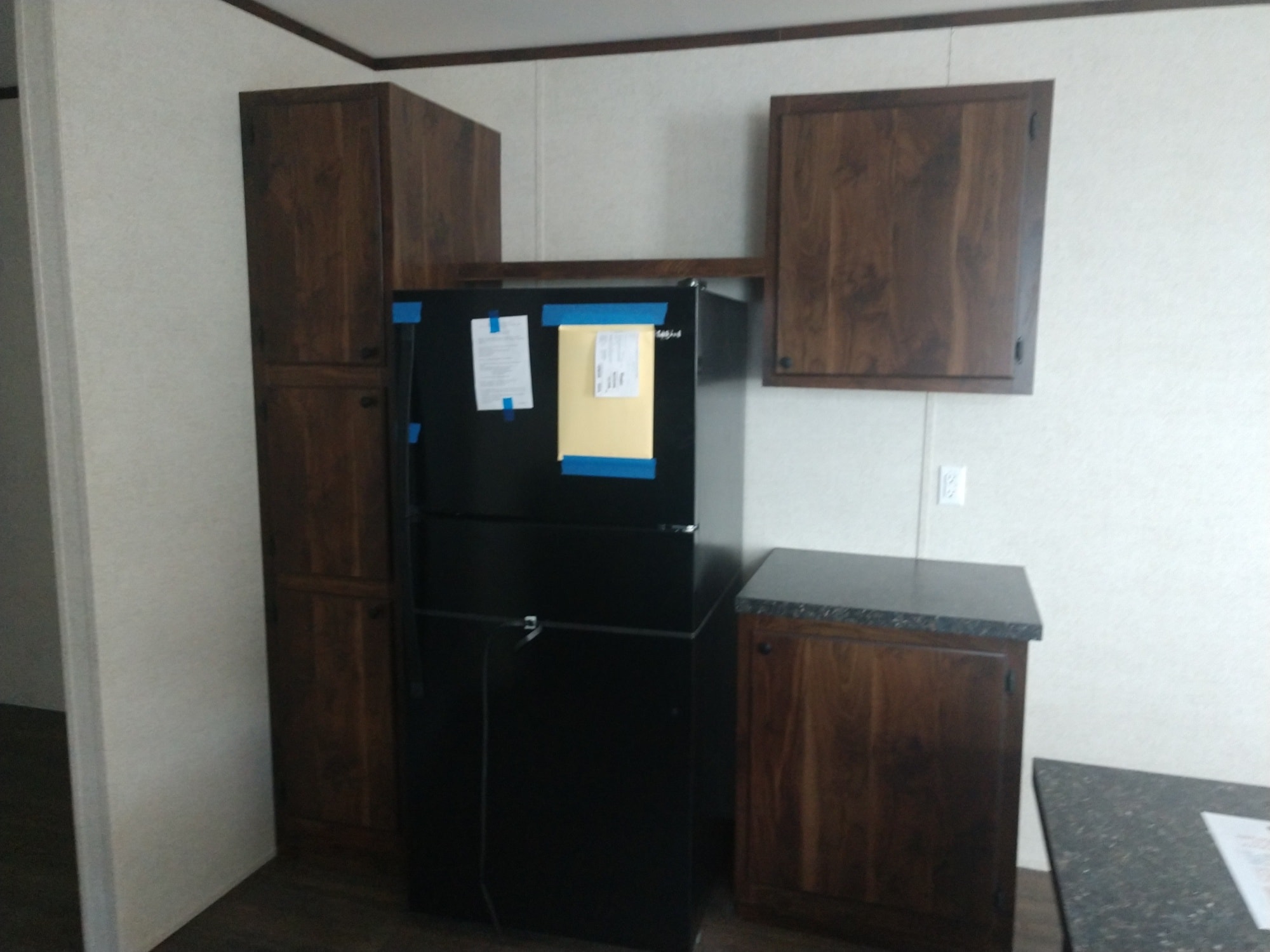 Kitchen with dark wood cabinets and a new black refrigerator, partially wrapped with blue tape. Minimalistic and clean ambiance with a neutral tone.