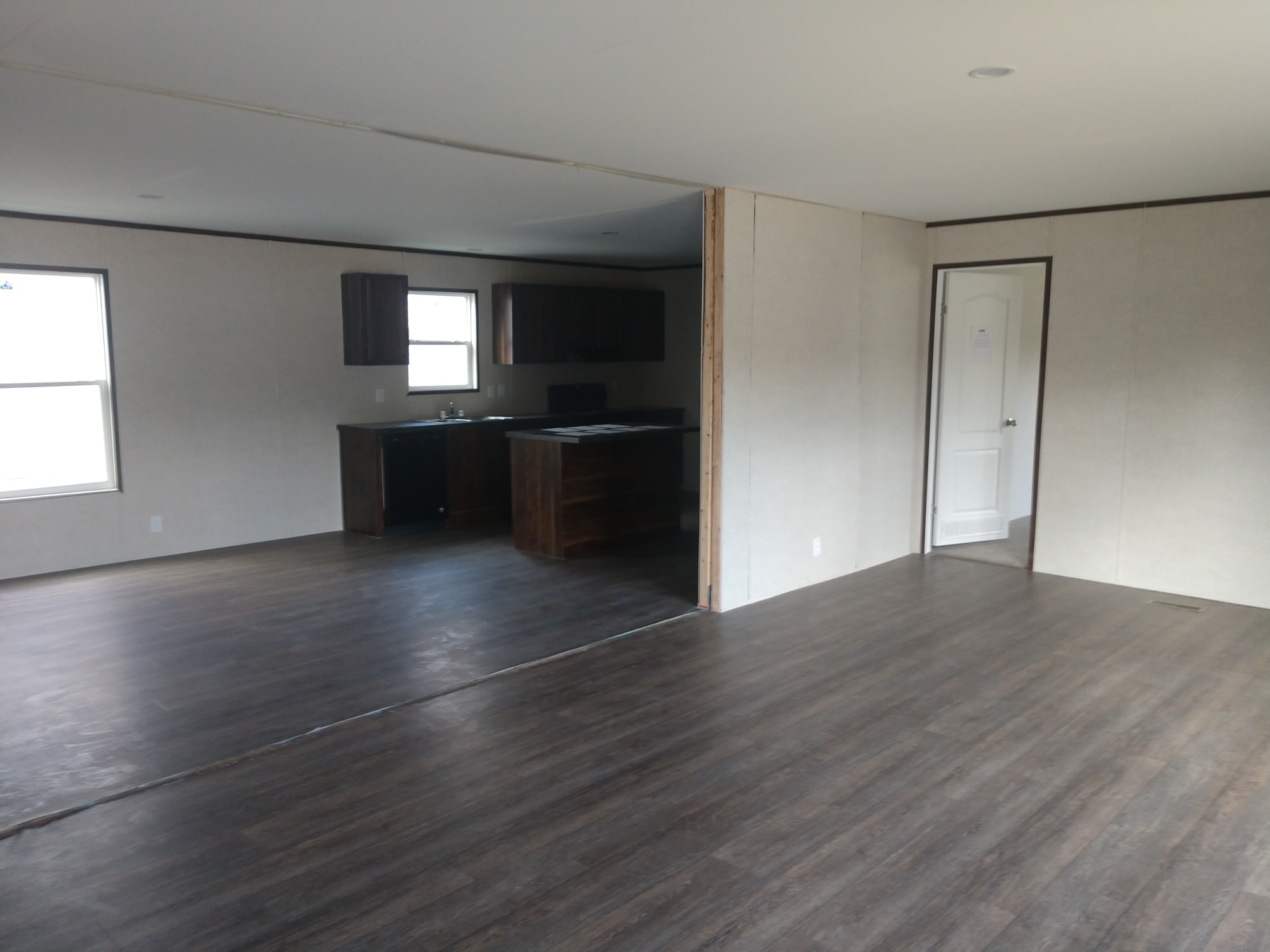 Spacious empty living area with dark wooden flooring, beige walls, and a view into a simple kitchen with dark cabinets. Minimalist and modern.