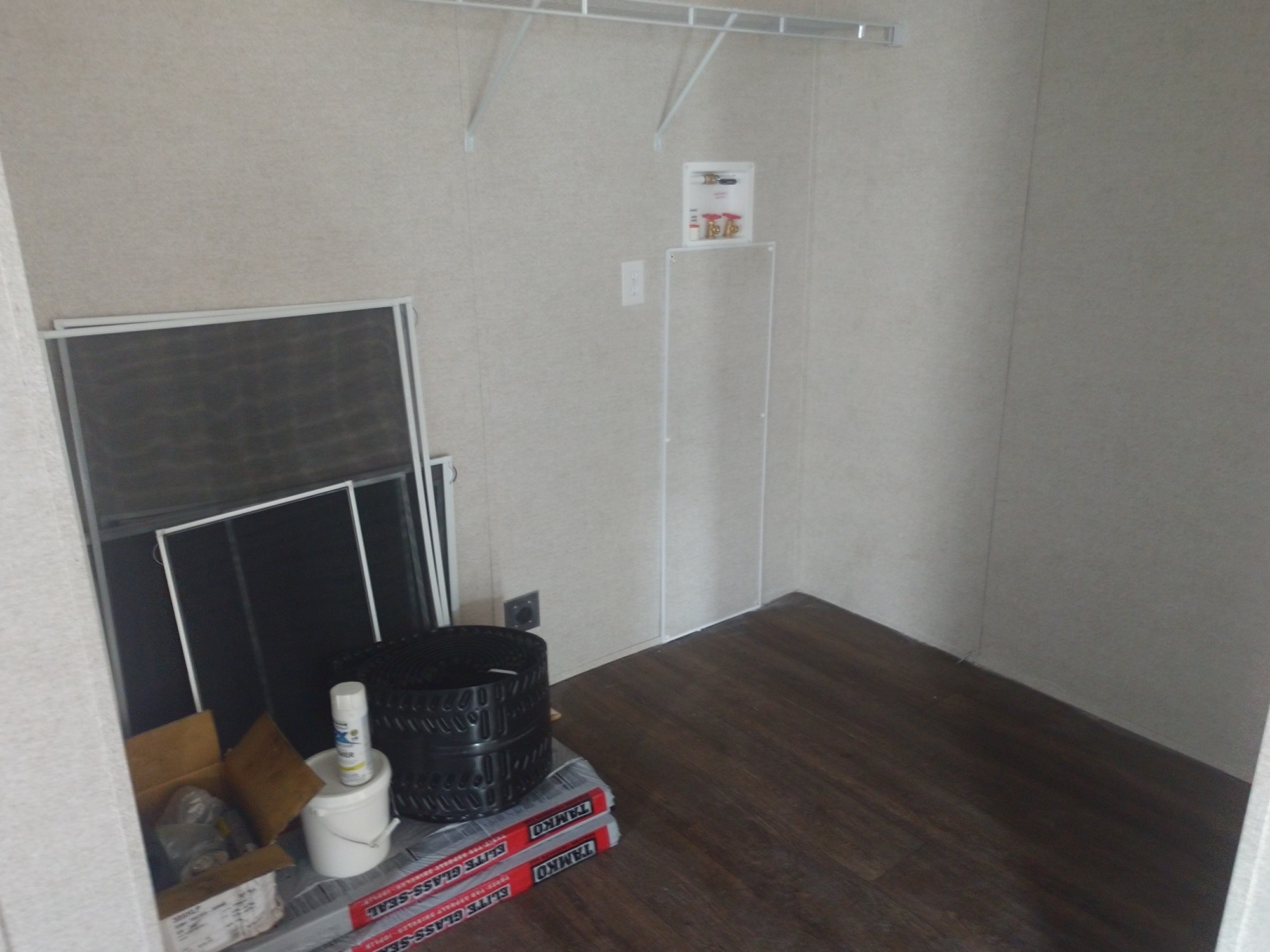 Small utility space with a wooden floor, beige walls, and metal shelving above. It contains window screens, tires, and cleaning supplies, conveying a storage function.