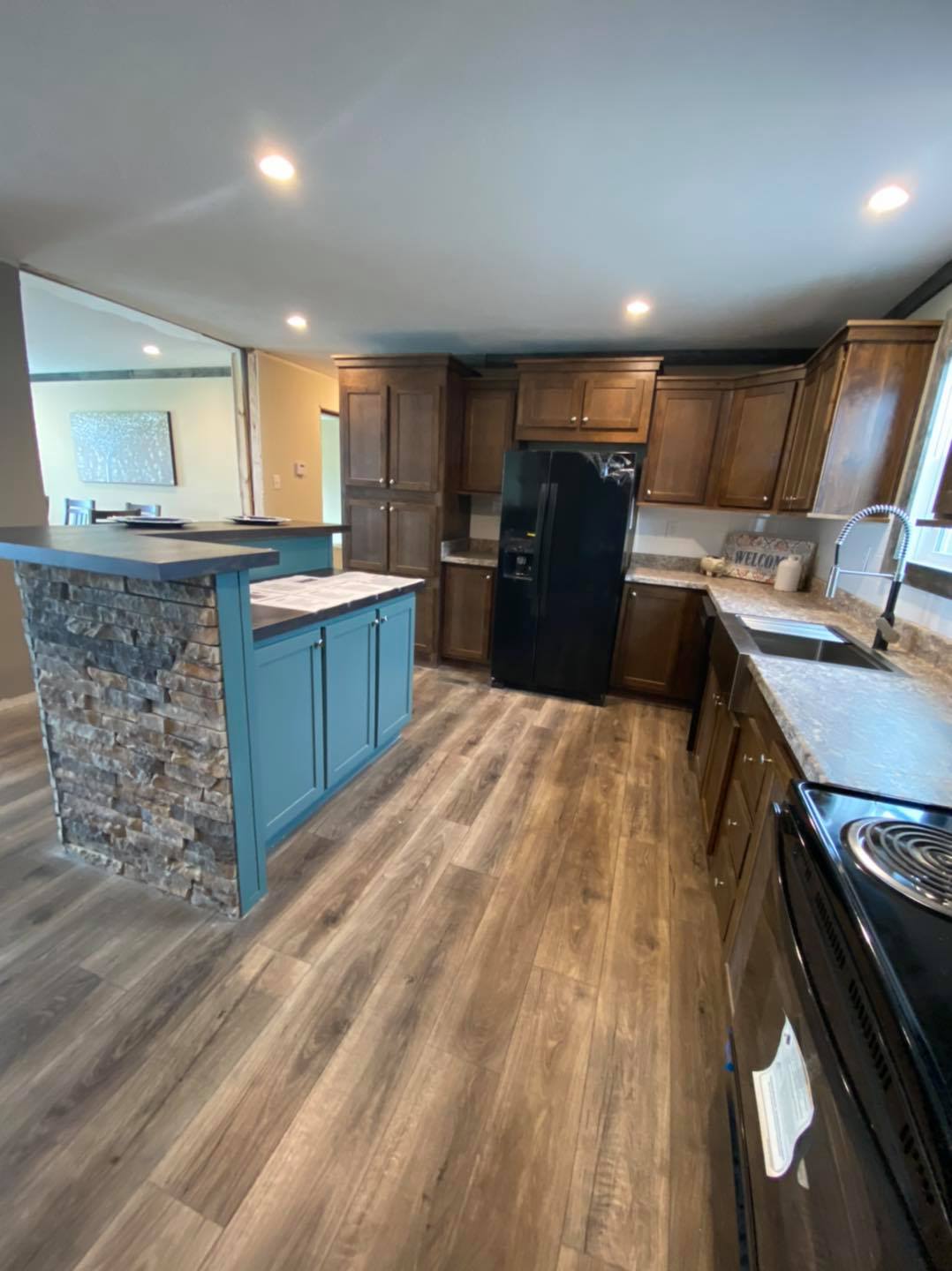 Modern kitchen with wood floors, dark wooden cabinets, and a stone island with teal accents. Includes a black fridge and stainless steel sink.