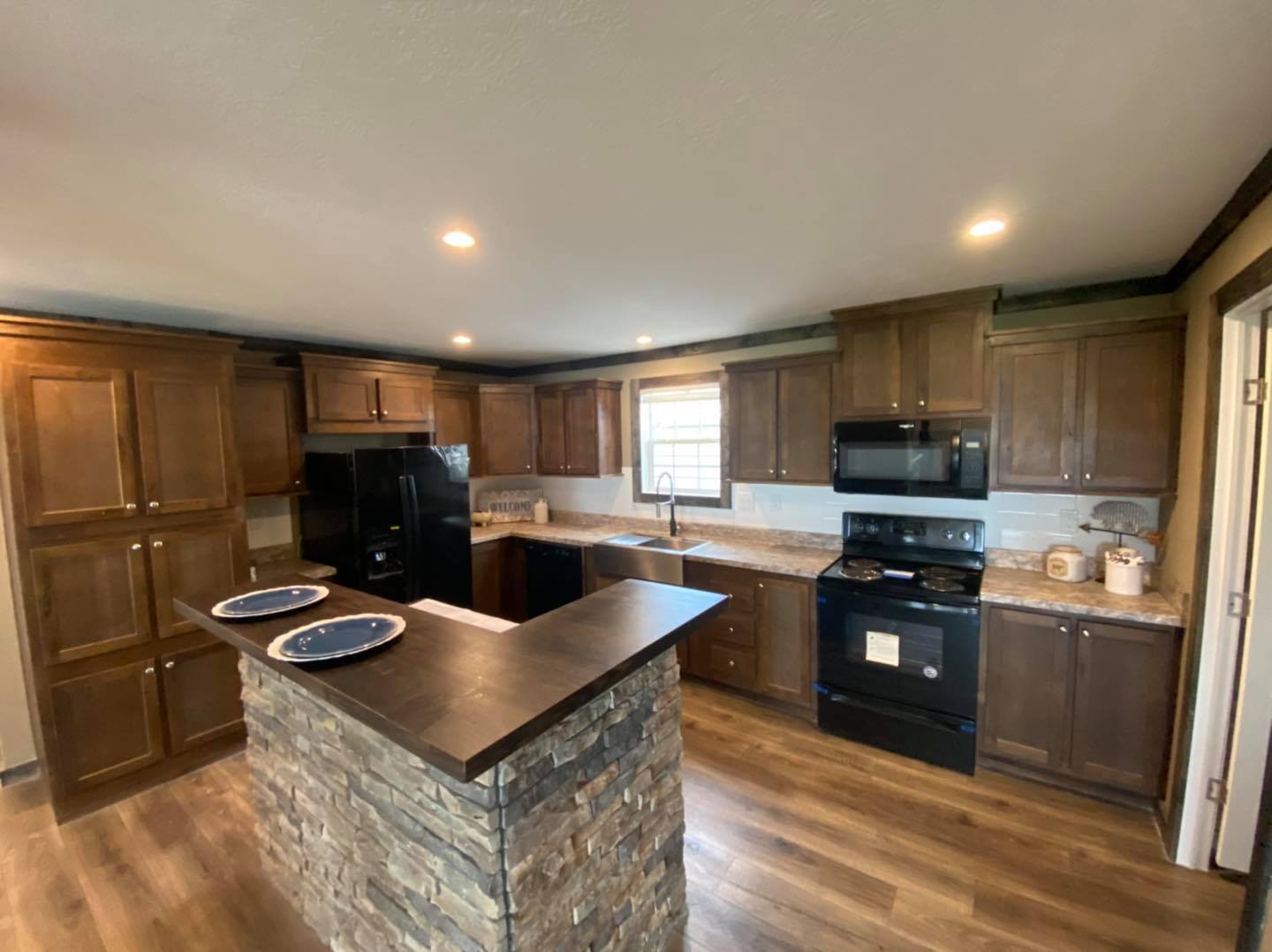 Spacious kitchen with wooden cabinets, a black fridge, stove, and microwave. Stone island with dark countertop, two plates, and warm lighting. Cozy atmosphere.