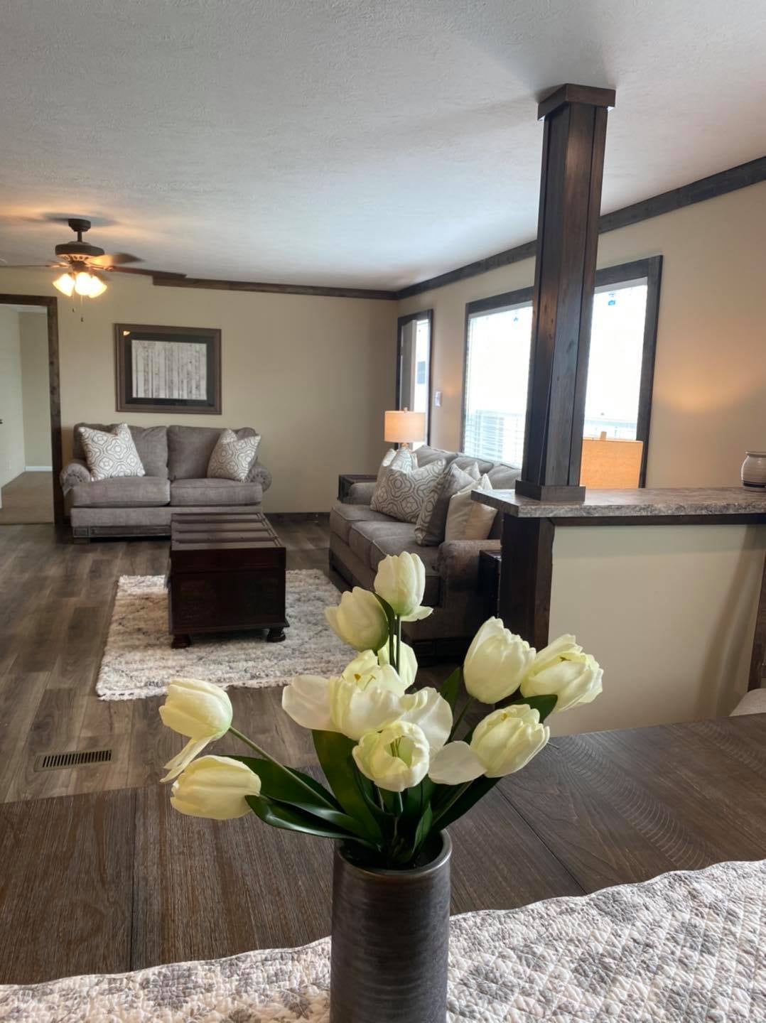 Cozy living room with gray sofas, decorative pillows, wooden coffee table, and warm lighting. White tulips in the foreground add a fresh touch.