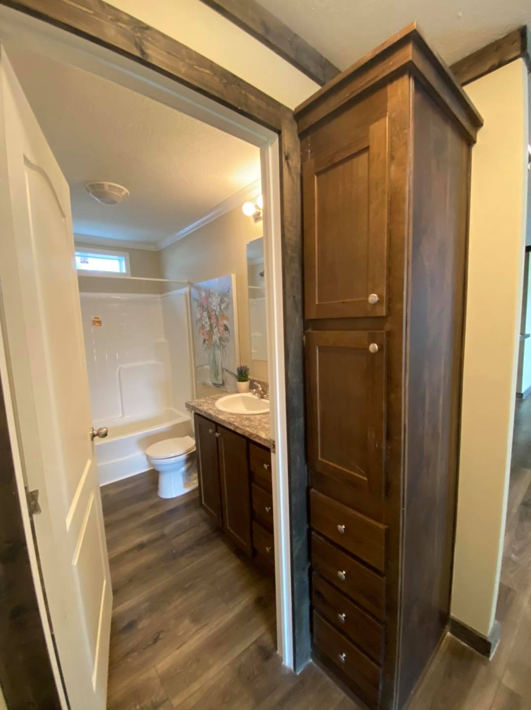 Warm, inviting bathroom with wood flooring, dark wooden cabinets, and a granite countertop. Features a white bathtub and a small, colorful wall art.