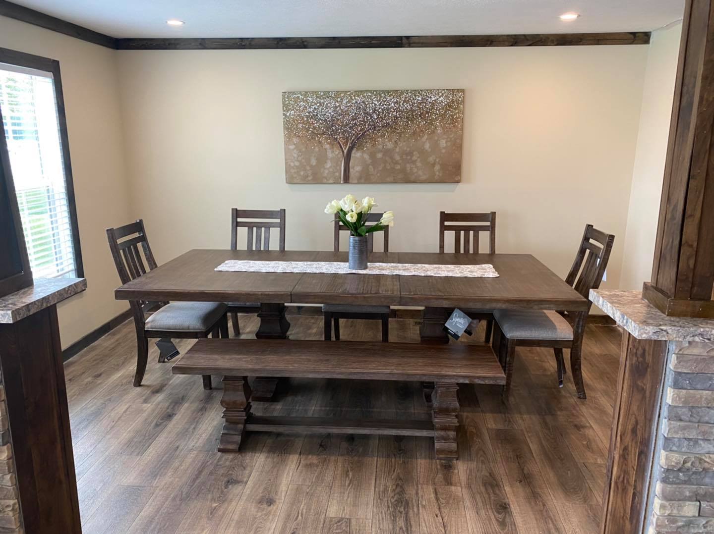 Dining room with a dark wooden table, matching chairs, and a bench. A vase with white flowers sits on a runner. Soft lighting and tree artwork create a cozy atmosphere.