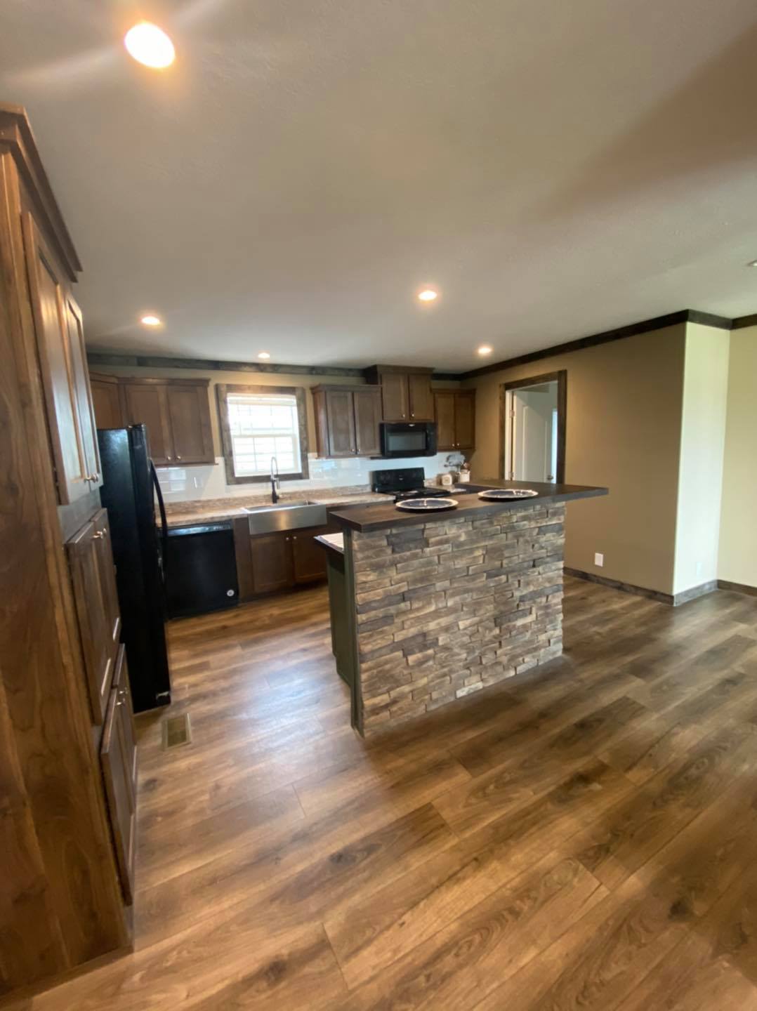 Modern kitchen with wooden flooring, brown cabinets, and a stone-accented island. Includes black appliances and warm lighting, creating a cozy atmosphere.