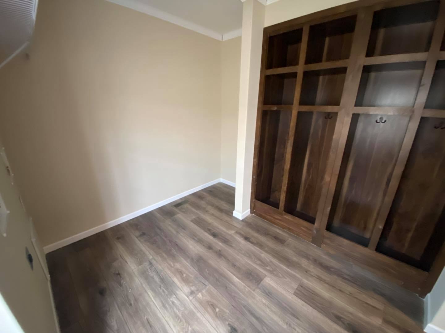 Empty room with beige walls and wood flooring, featuring a large wooden built-in wardrobe on the right. The room is simple and unfurnished.