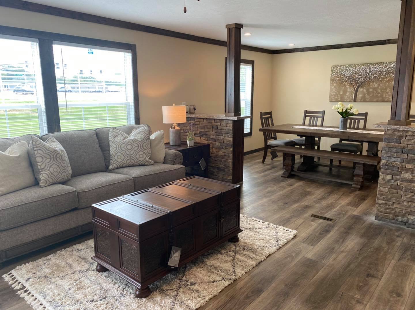 Cozy living room with a gray sofa and decorative pillows, a wooden chest as a coffee table on a textured rug, and a dining area with a wooden table and chairs. Warm, welcoming ambiance.