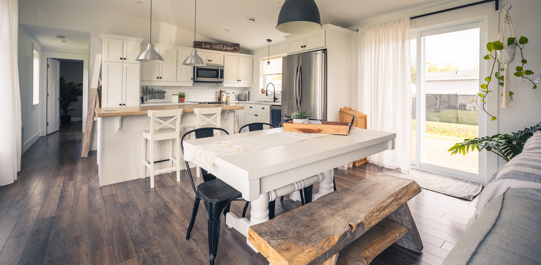 A bright, modern kitchen and dining area with white cabinetry, wooden floors, a table with mixed seating, pendant lights, and a sliding glass door.