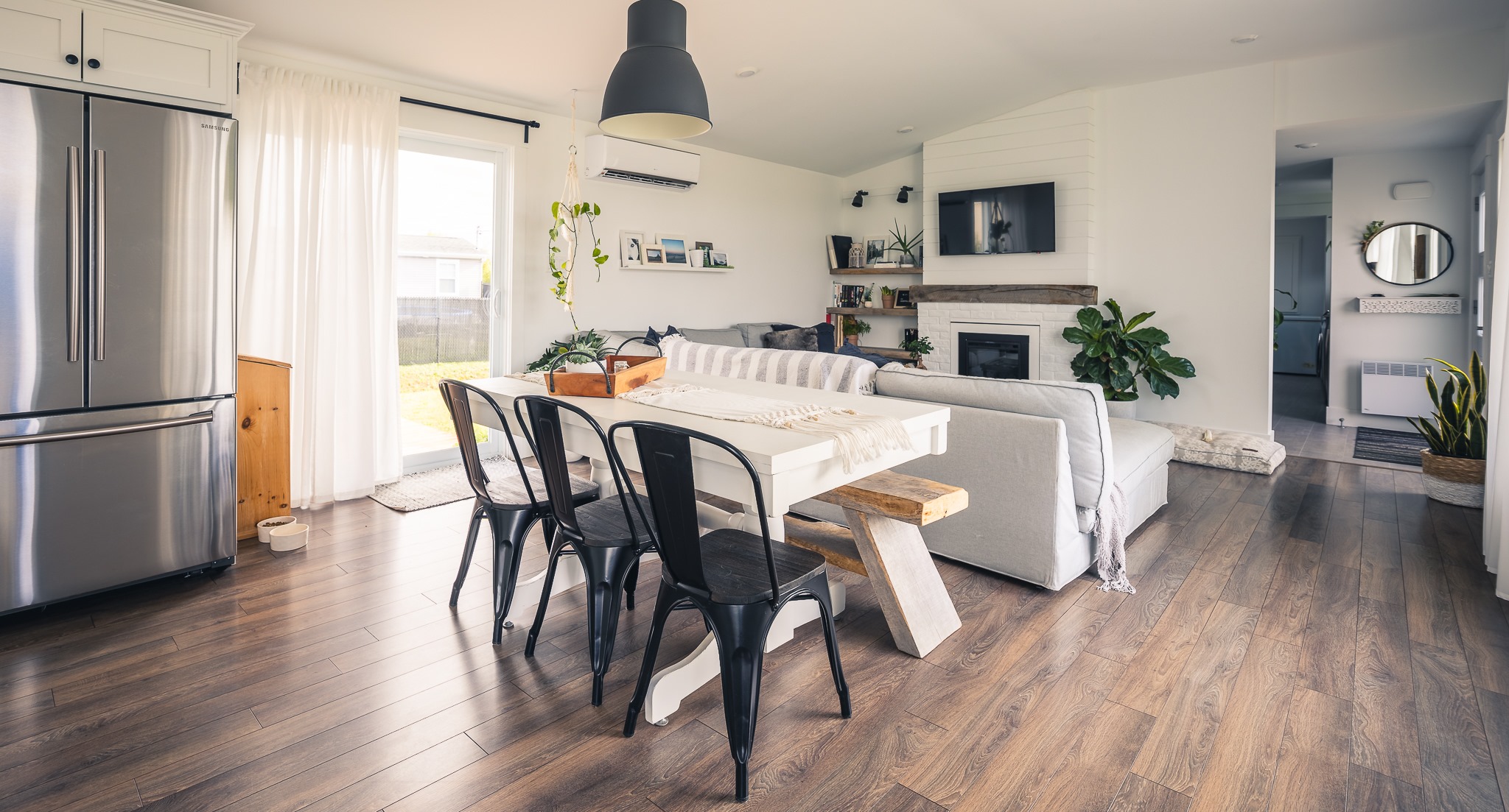 A modern, bright living and dining area with wooden floors and white walls. Features a table with black chairs, a sofa, fireplace, and large windows. Cozy and inviting.