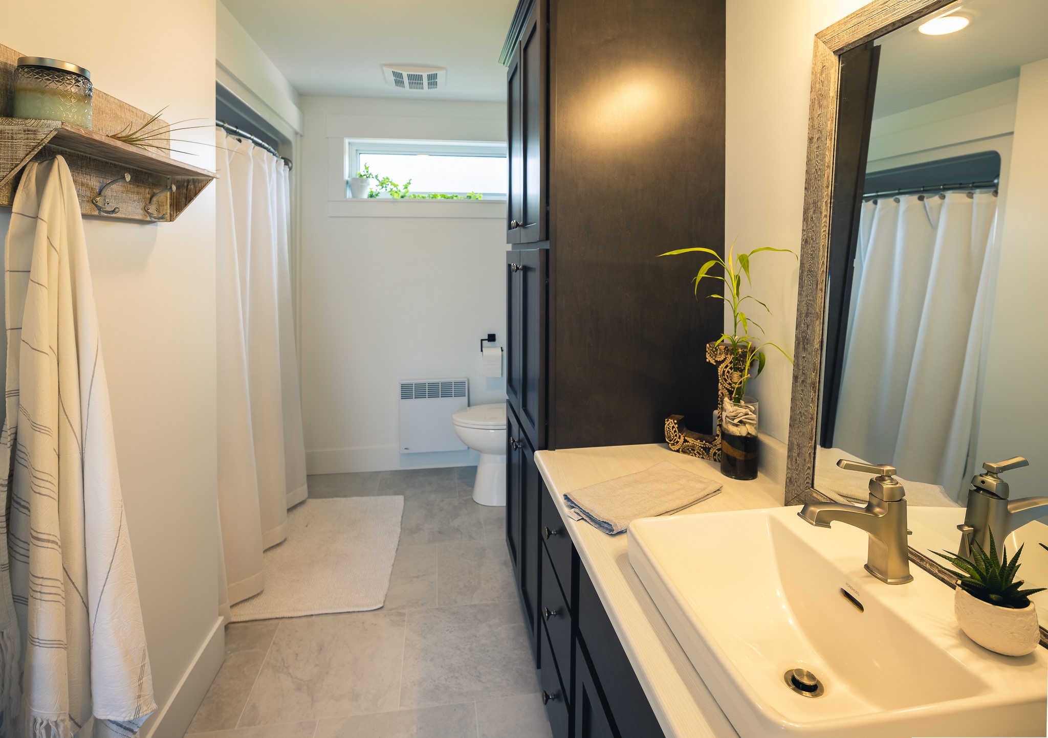 Modern bathroom with a beige color scheme, featuring a white sink, dark cabinetry, potted plants, and a towel on a rack. Natural light enters through a small window above the toilet, creating a clean and serene atmosphere.