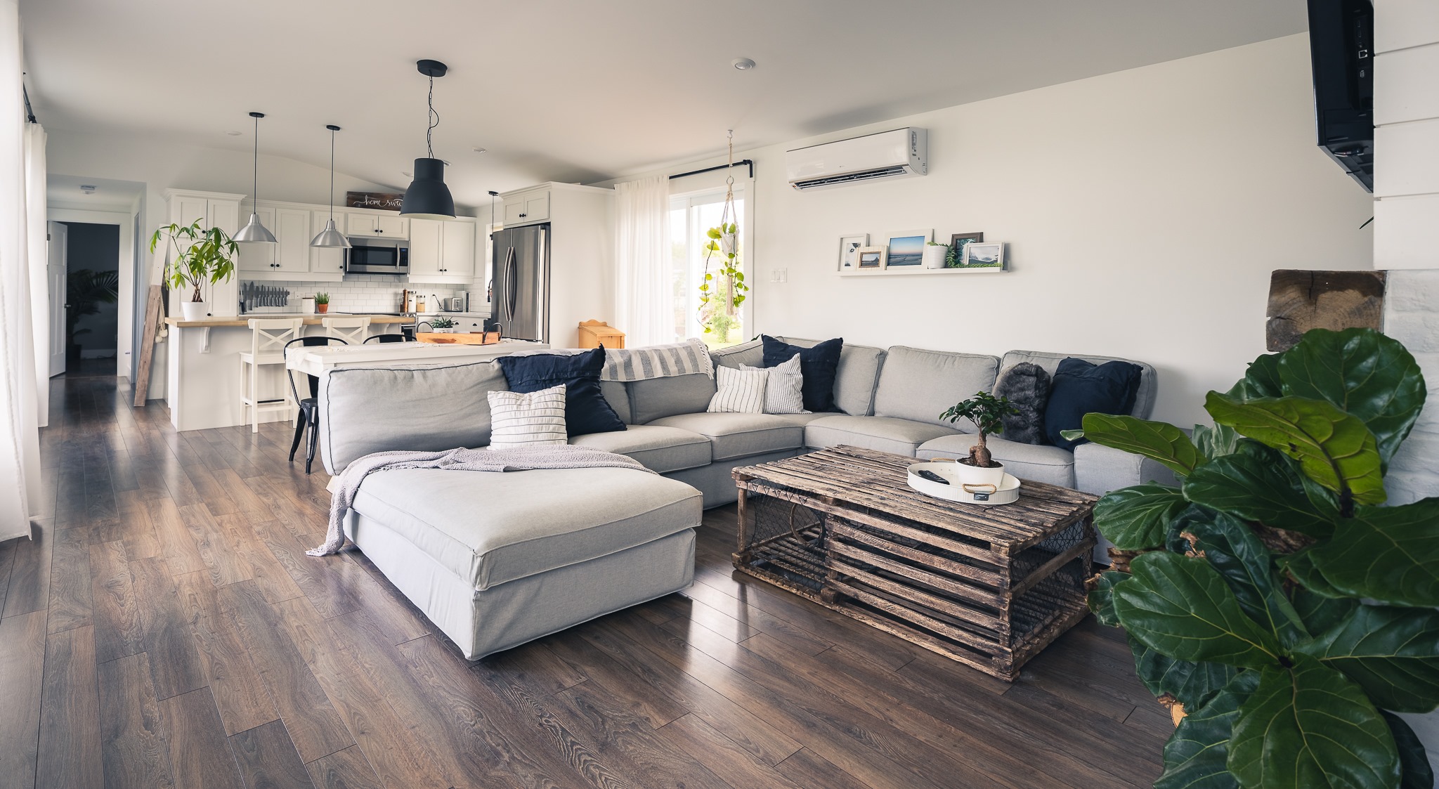 A cozy, modern living room with a large gray sectional sofa, wooden coffee table, and plants. The background features a bright kitchen with white cabinets.