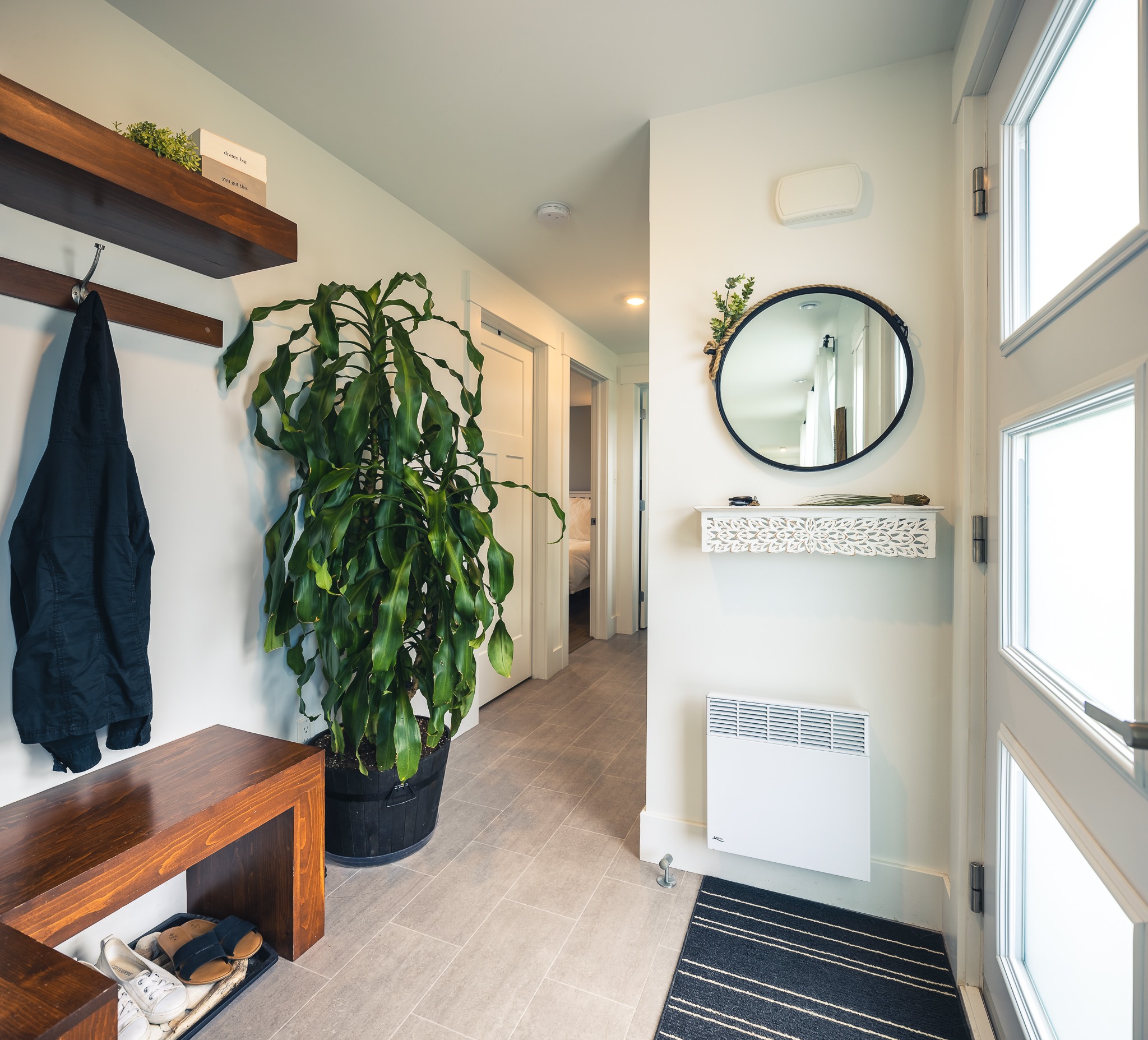Modern entryway with a large potted plant, wooden bench, coat hook, and shoes. A round mirror and a shelf enhance the bright, welcoming space.
