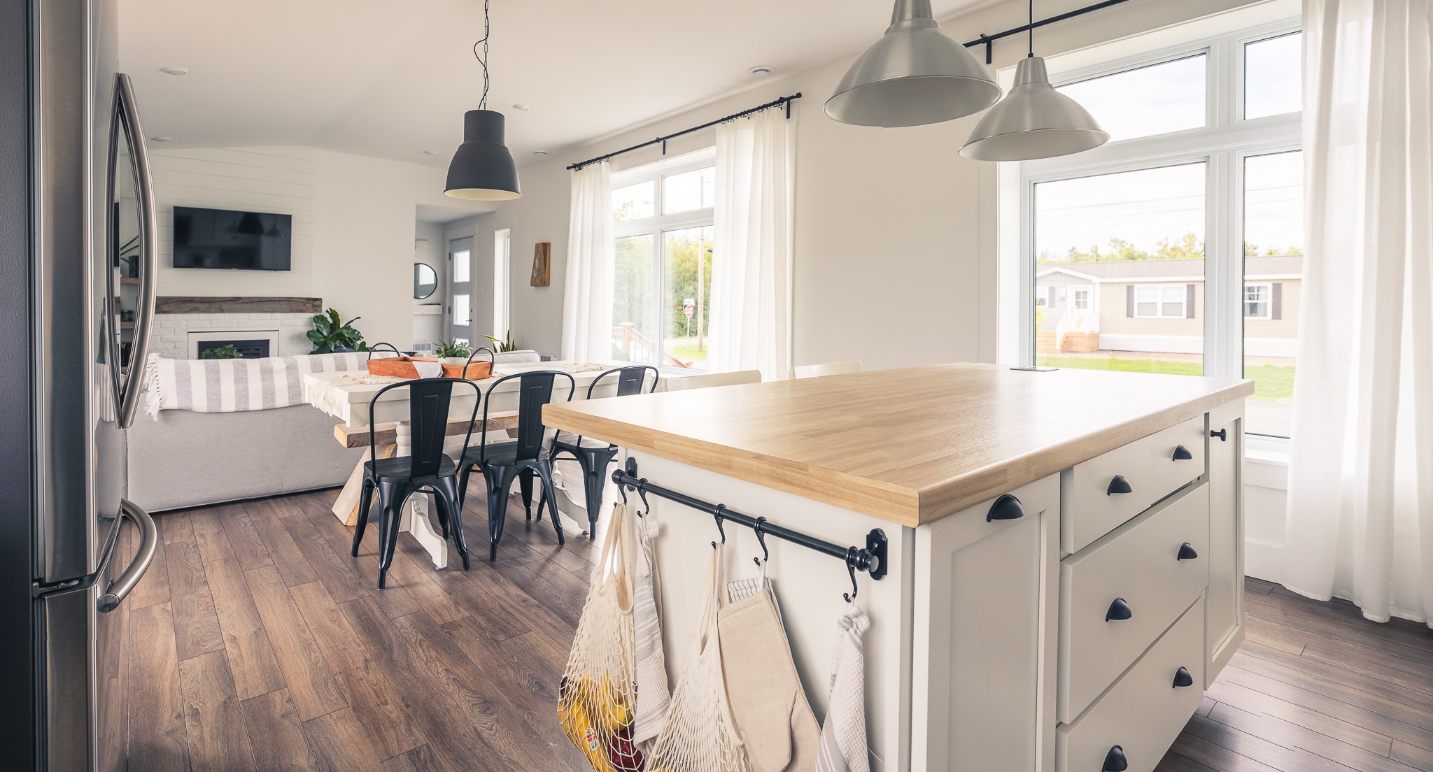 A bright, modern kitchen and dining area with wooden floors, an island with a wooden countertop, pendant lights, and a cozy living room in the background.