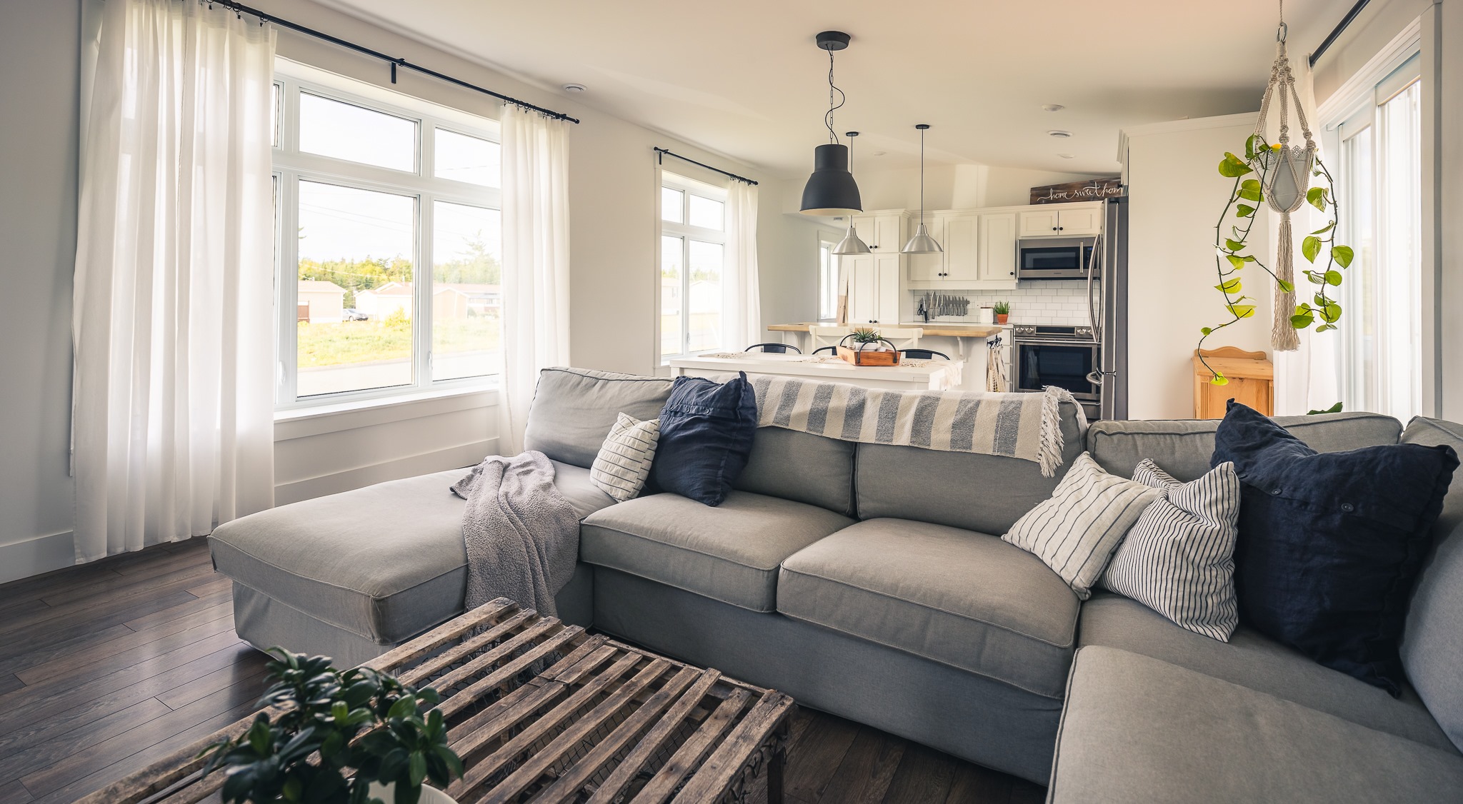 Spacious, sunlit living room with a gray sectional sofa showcasing blue and white pillows. A cozy, inviting atmosphere with a view of a modern kitchen.