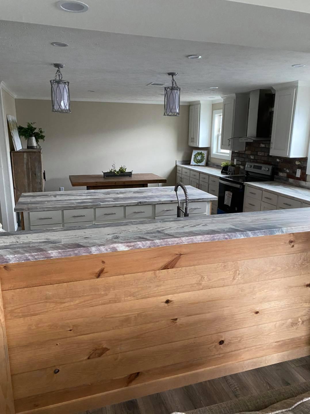 Spacious kitchen with wooden and gray accents, light fixtures, and a distant dining table. Includes modern appliances and a brick backsplash, creating a cozy, rustic feel.