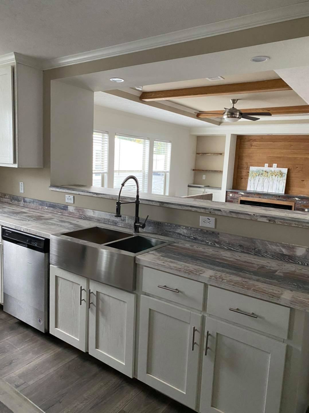 Modern kitchen with white cabinets, stainless steel appliances, and a double farmhouse sink. A window opens to a bright living room with ceiling fan.