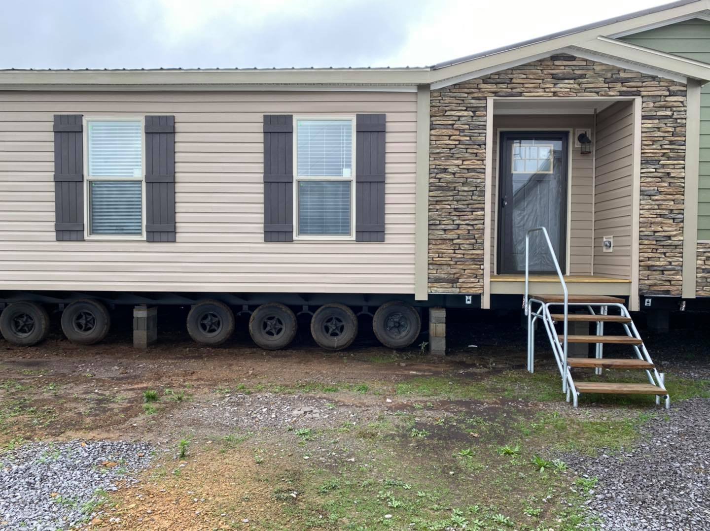 A mobile home on wheels with a beige siding exterior, dark shutters, and stone accent. Metal stairs lead to the entrance, set against a gravel and grass ground.