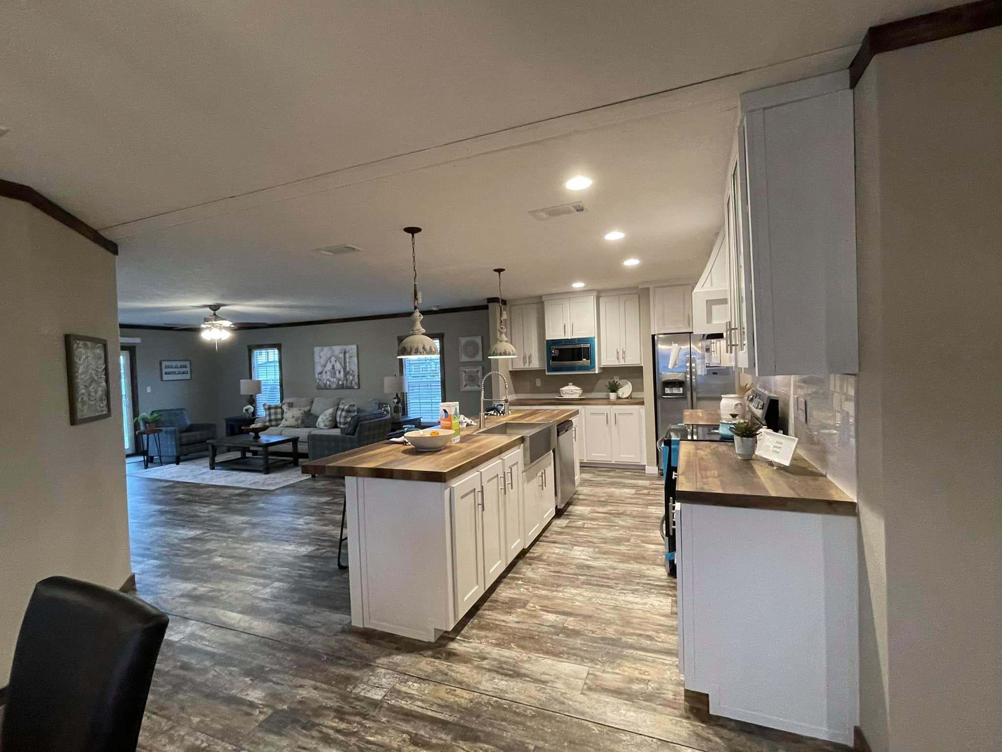 Spacious kitchen with white cabinets and wood countertops, center island with pendant lights. Open living area with sofas in the background. Warm, inviting tone.