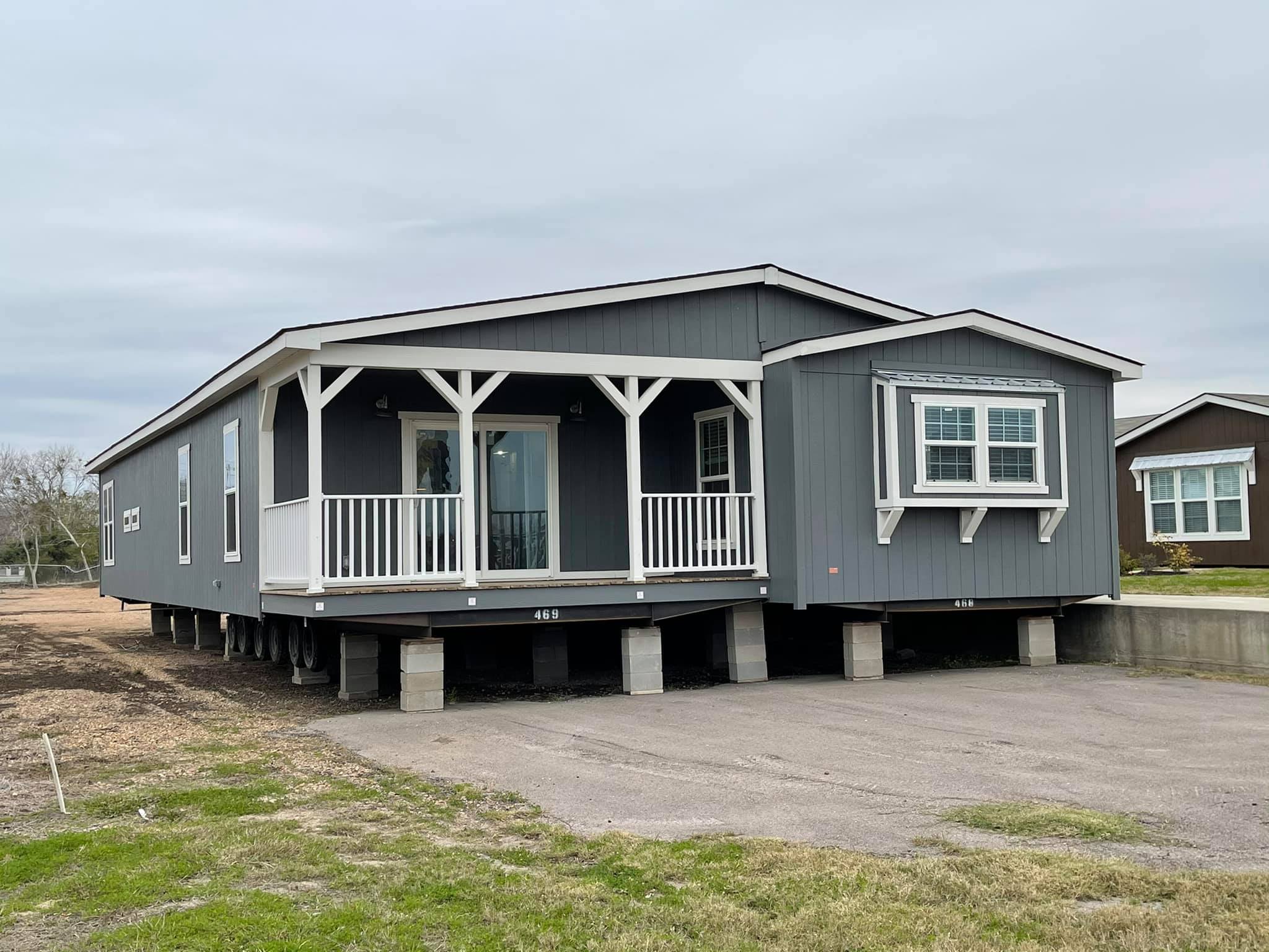 Grey manufactured home with white trim, featuring a small covered porch on the left. Set on a stone foundation, surrounded by a grassy yard and bushes.
