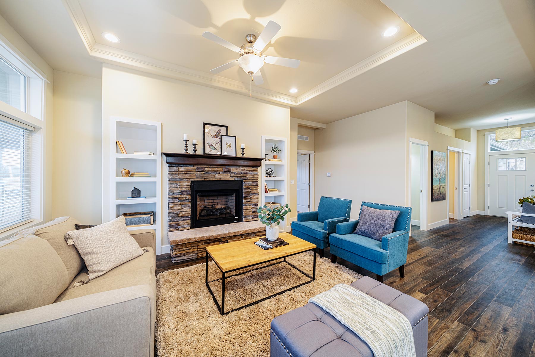 Modern living room with a cozy ambiance, featuring a beige sofa, two blue armchairs, a stone fireplace, and wood floors. Natural light filters through large windows.