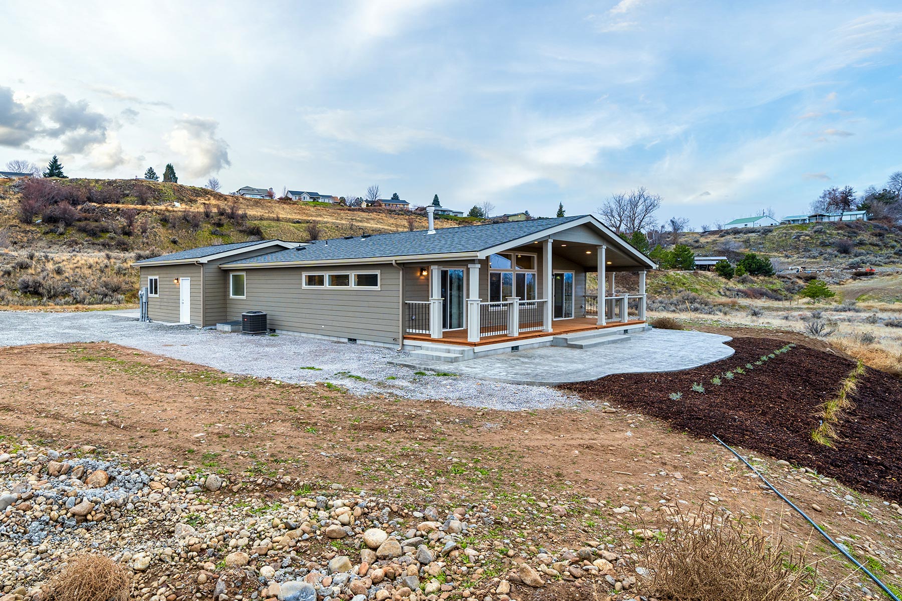 A modern, single-story house with a wraparound porch set in a hilly landscape. The home is surrounded by rocky ground and sparse vegetation under a partly cloudy sky.