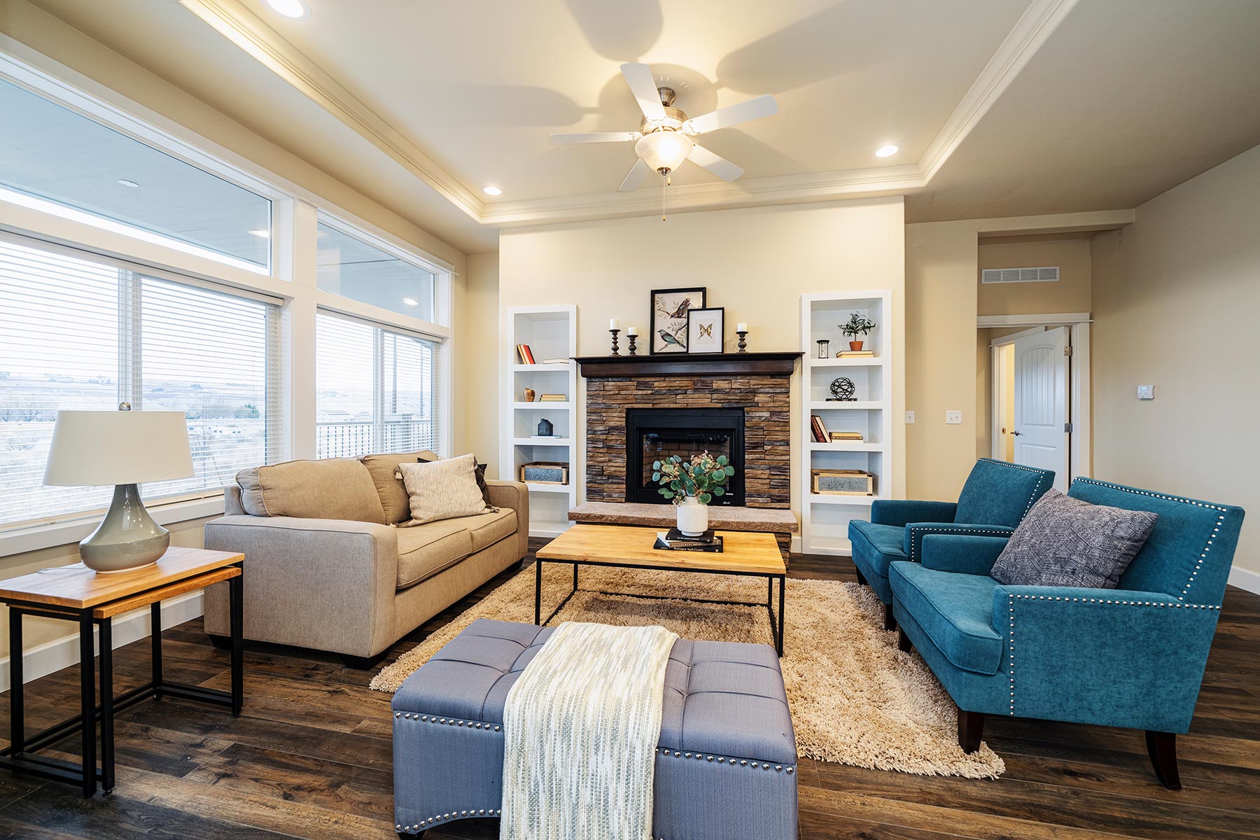 Cozy living room with a beige sofa, two teal armchairs, and a wooden coffee table. Stone fireplace, large windows, and warm lighting create a welcoming atmosphere.