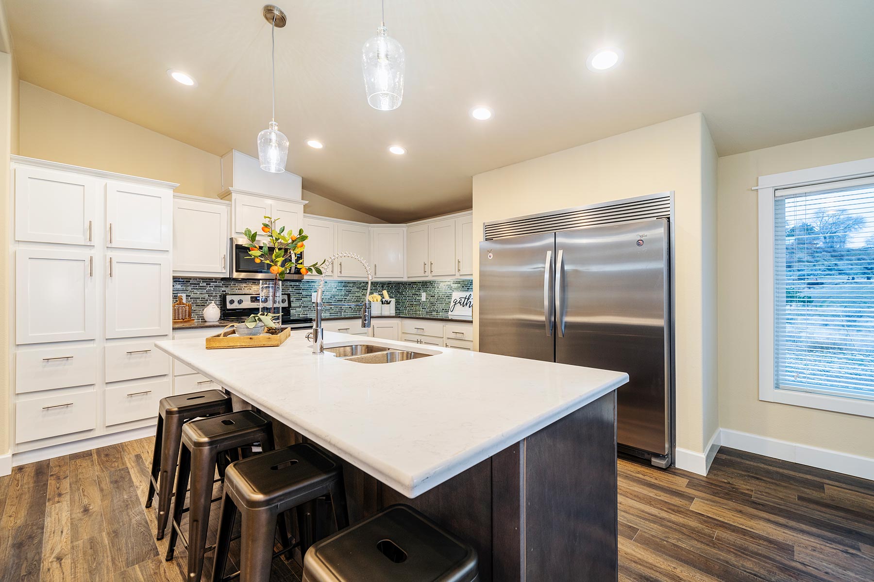Modern kitchen with white cabinets, dark wood flooring, and stainless steel appliances. Center island features a sink and black stools beneath elegant lighting.