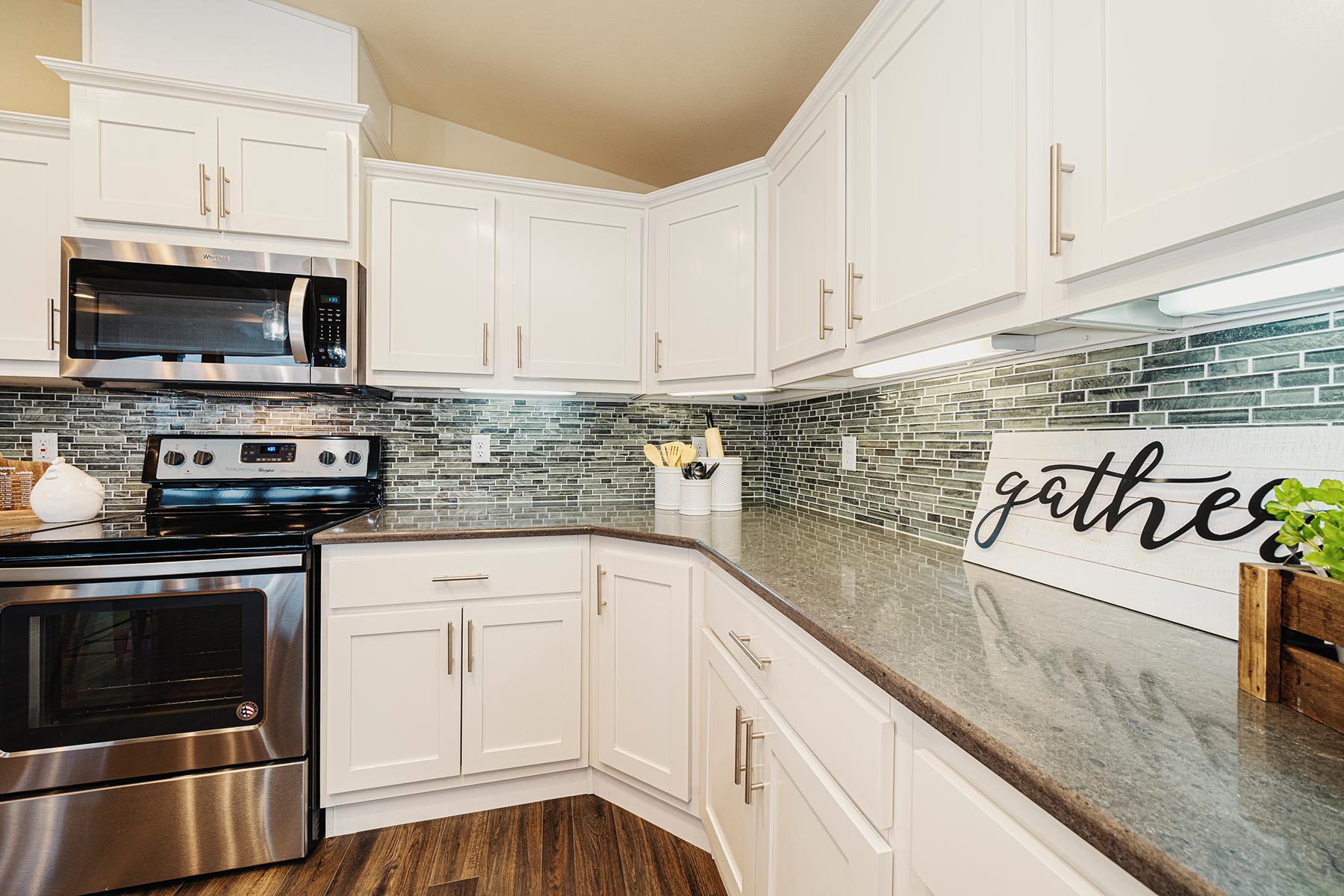 Modern kitchen with white cabinets, stainless steel appliances, and a gray tiled backsplash. A "gather" sign and plant add warmth. Wood floor.