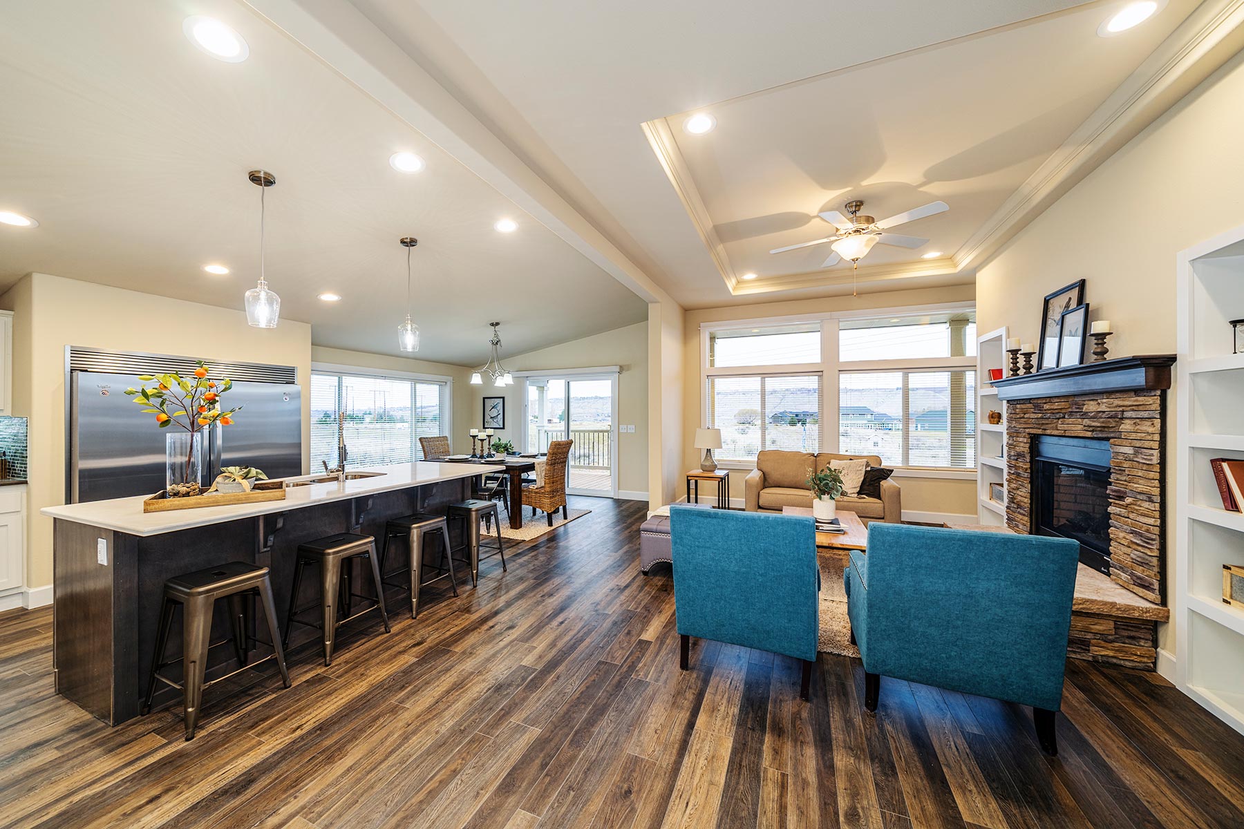 Spacious living room with wood flooring, featuring blue armchairs, a stone fireplace, and large windows. An open kitchen with barstools and pendant lights.