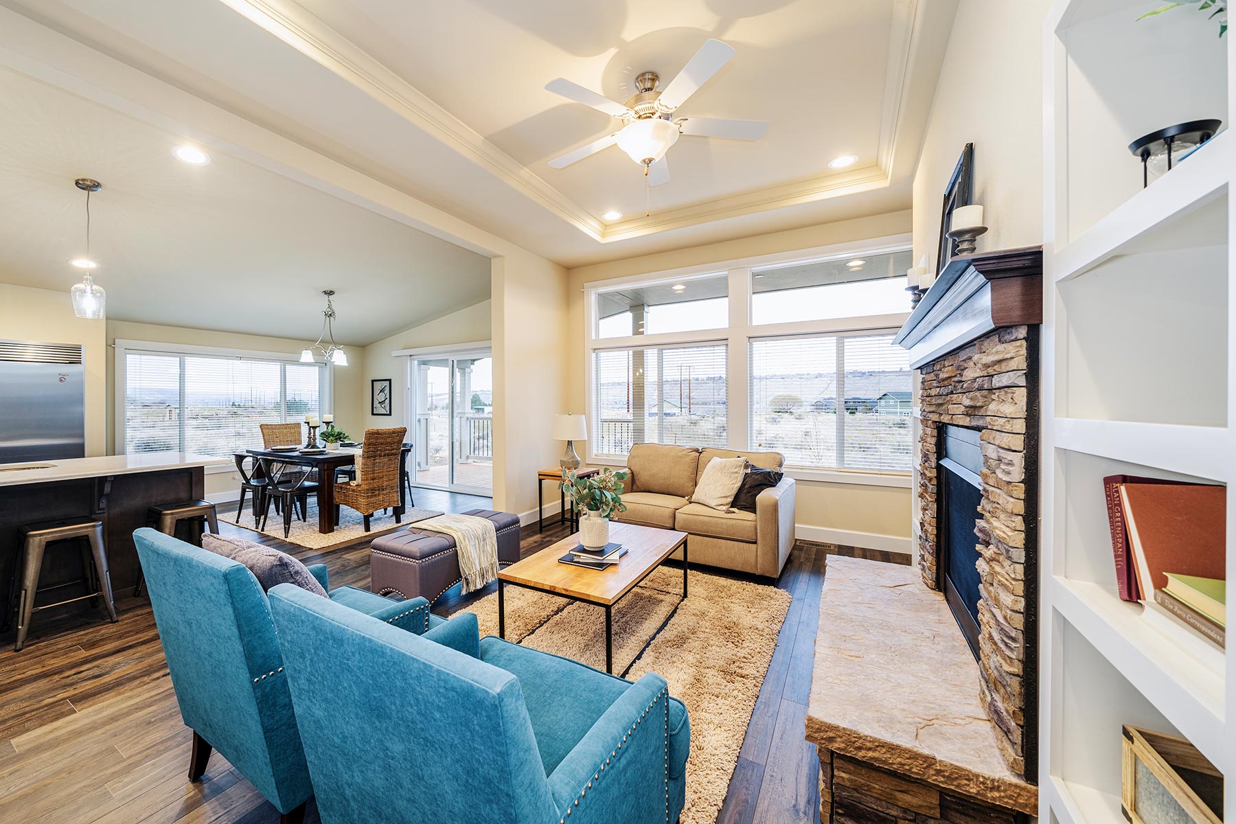 Bright, open living area with a stone fireplace, beige sofa, blue armchairs, and a wooden coffee table. Dining area and kitchen visible in background. Cozy and inviting.