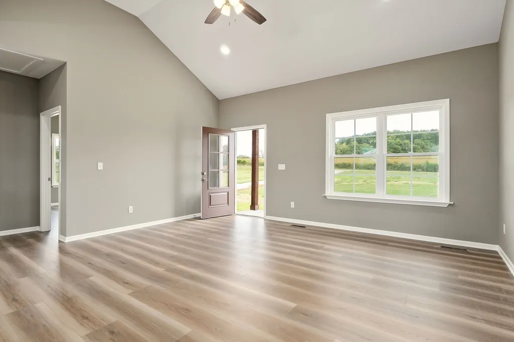 Spacious room with light wood flooring and soft gray walls. A large window and open door reveal a green outdoor scene under a bright ceiling fan. Calm ambiance.