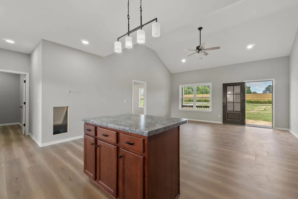Spacious modern kitchen and living area with gray walls, wood floors, and a central island. Large window and door reveal a sunny outdoor view.