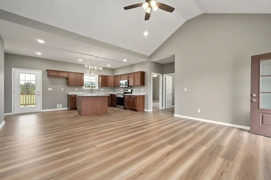 Spacious, modern kitchen and living area with vaulted ceiling, wooden floors, and ceiling fan. Features dark wood cabinets, island, and large windows.